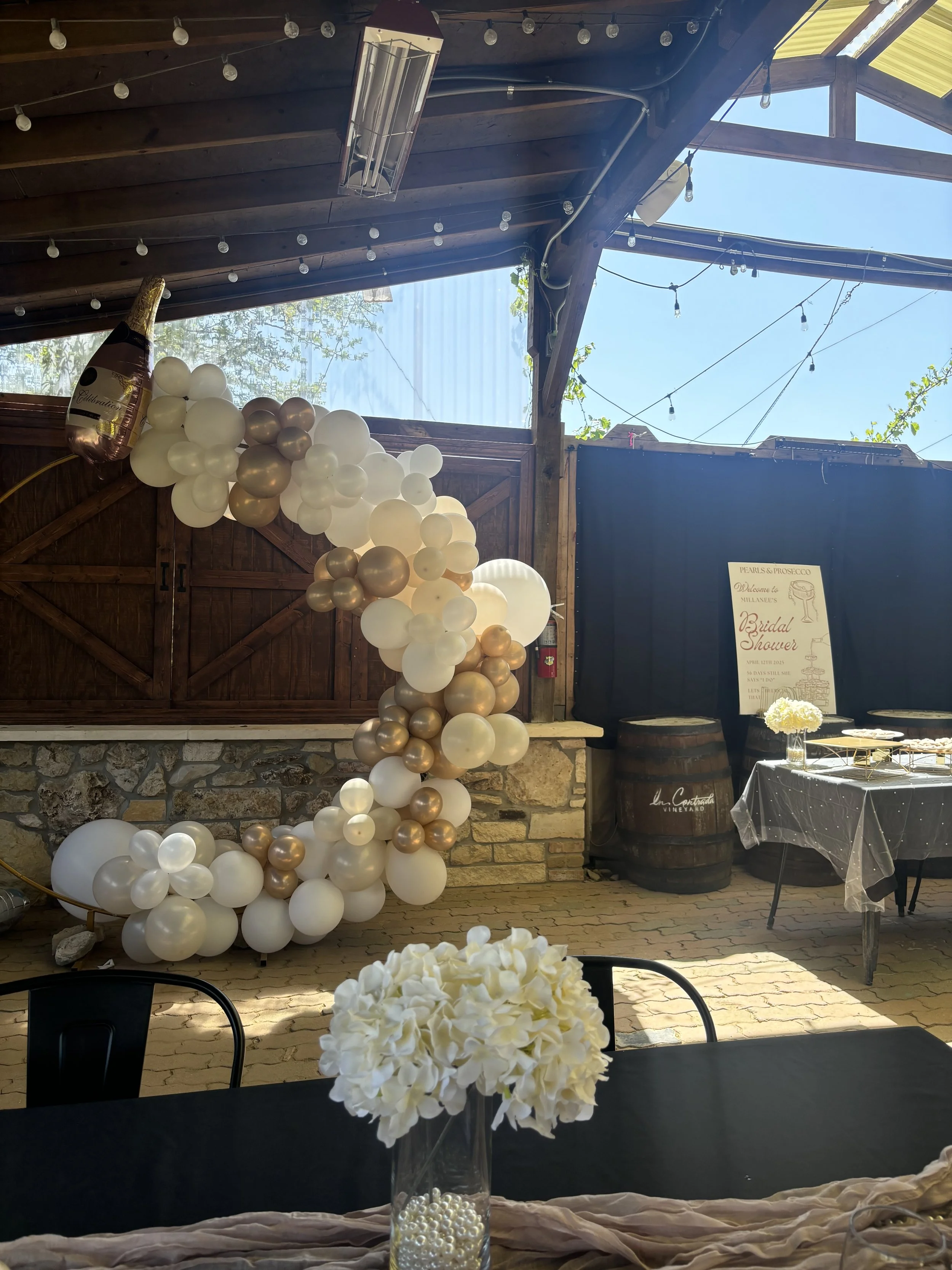 Decorative balloon arrangement with white and gold balloons at a bridal shower event, featuring a sign with event details and a white flower centerpiece on a table with a black tablecloth.