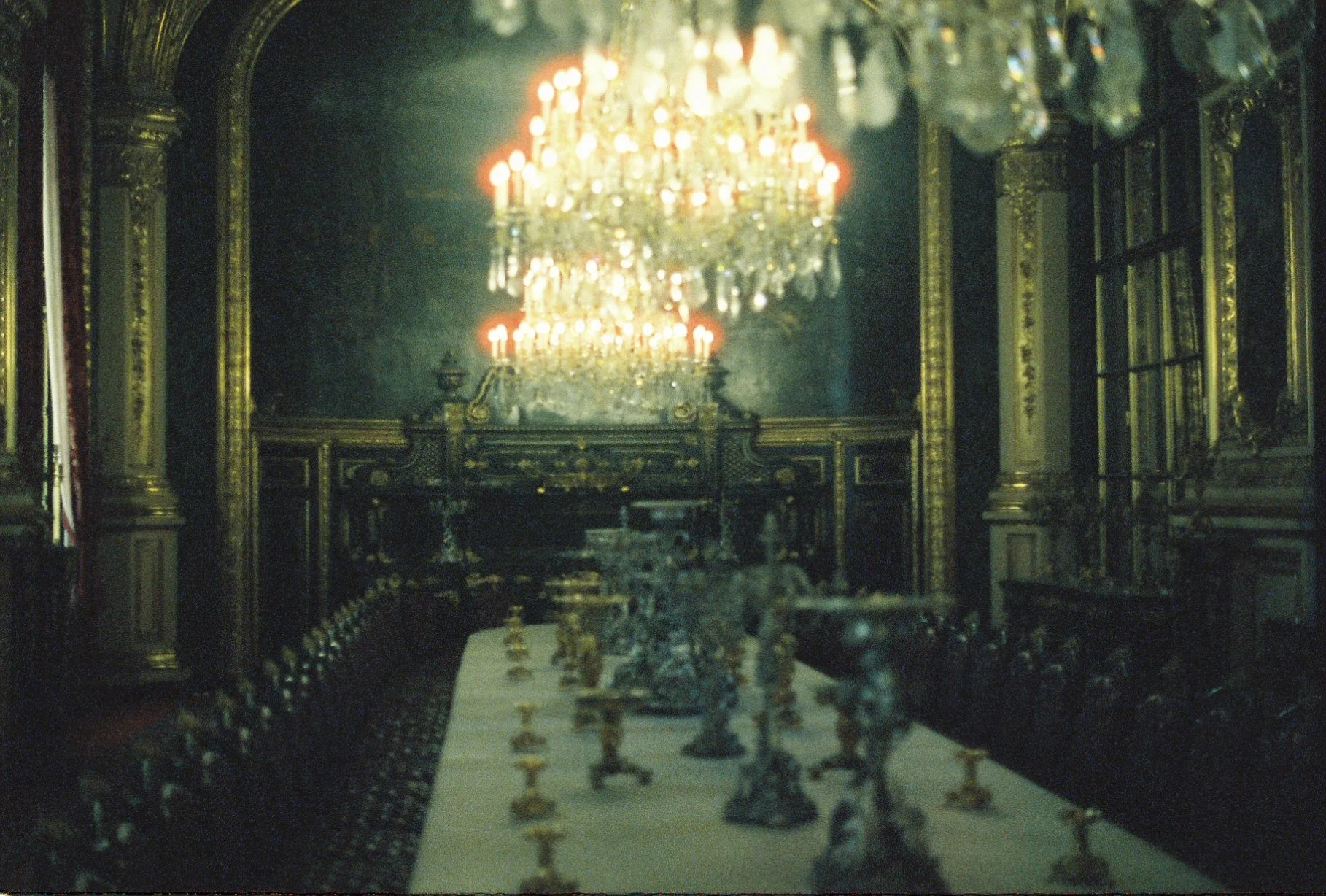 An opulent dining room with ornate gold-trimmed walls, a long table set with elaborate silverware and candles, a large chandelier hanging from the ceiling, and richly decorated chairs along the table, evoking a luxurious historical setting.