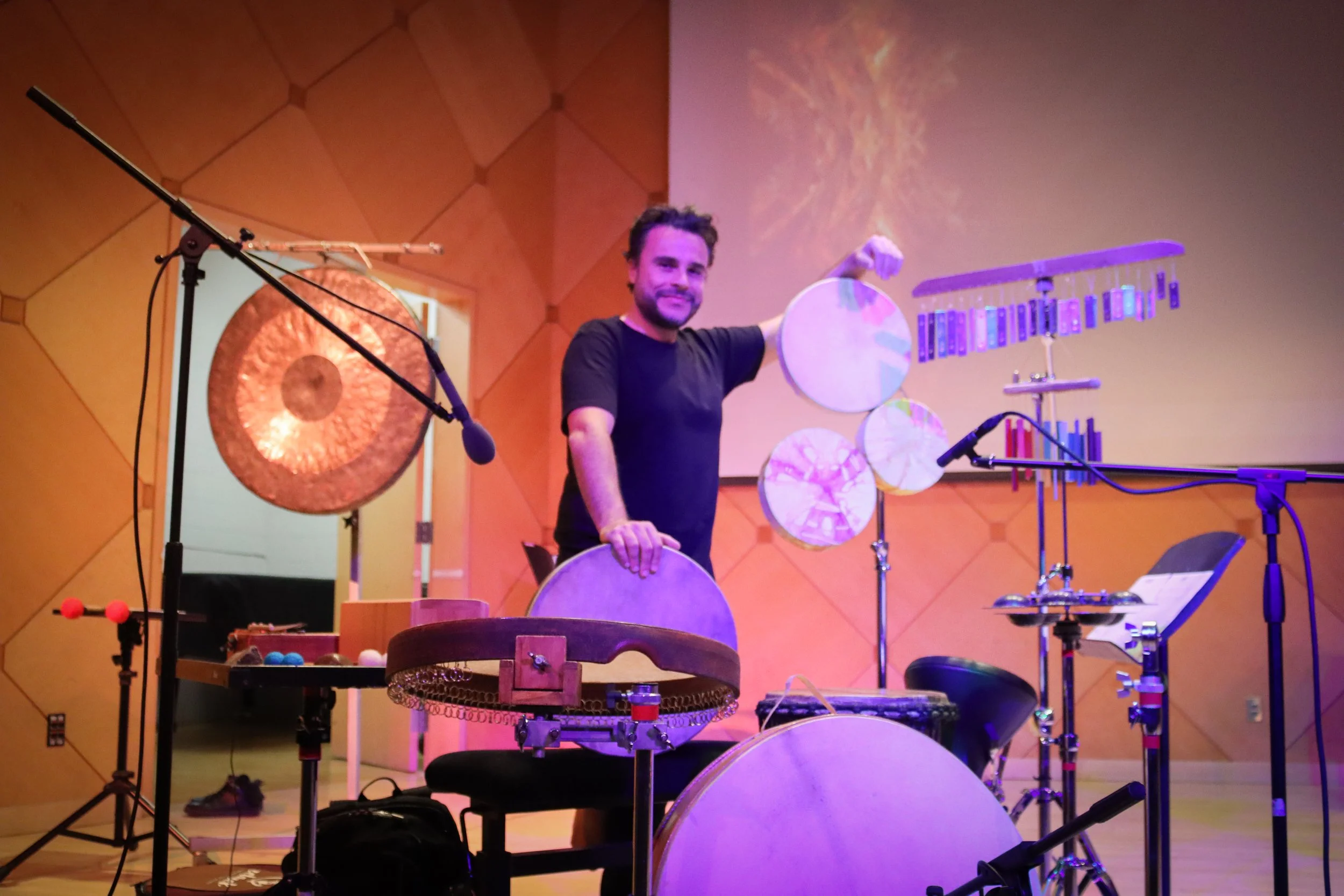 A man smiling on stage with percussion instruments, including xylophones and gongs, surrounded by microphones and music stands in a room with orange walls.