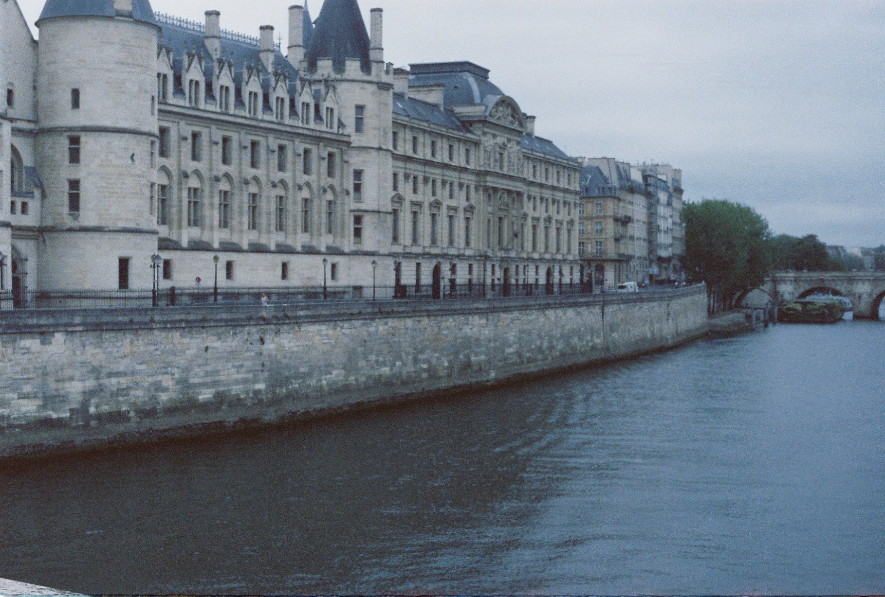 A row of historic European-style buildings along a river, with a stone bridge in the background and a tree on the right side of the riverbank.