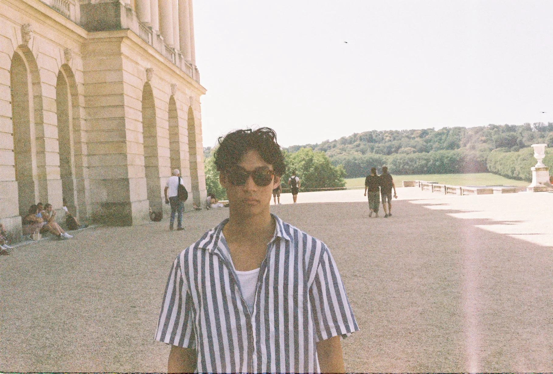 A young man with dark curly hair, wearing sunglasses and a striped shirt, standing outdoors near a historic building with arched windows and a landscape of trees and green hills in the background.