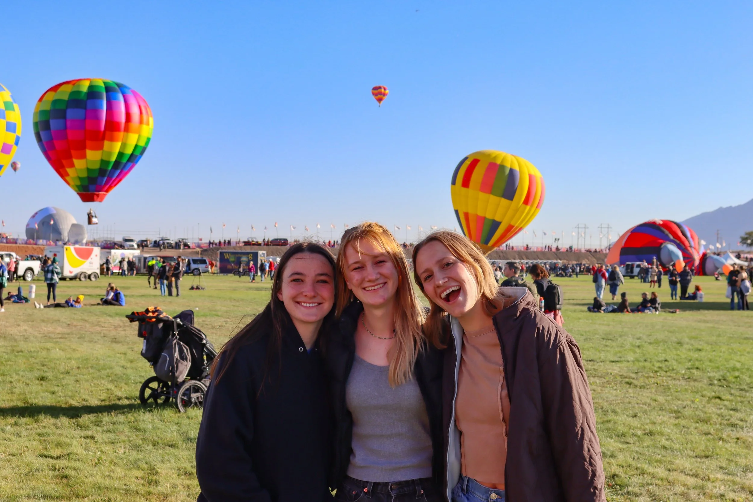 A group of three smiling young women standing together outdoors at a hot air balloon festival with colorful hot air balloons in the sky behind them.