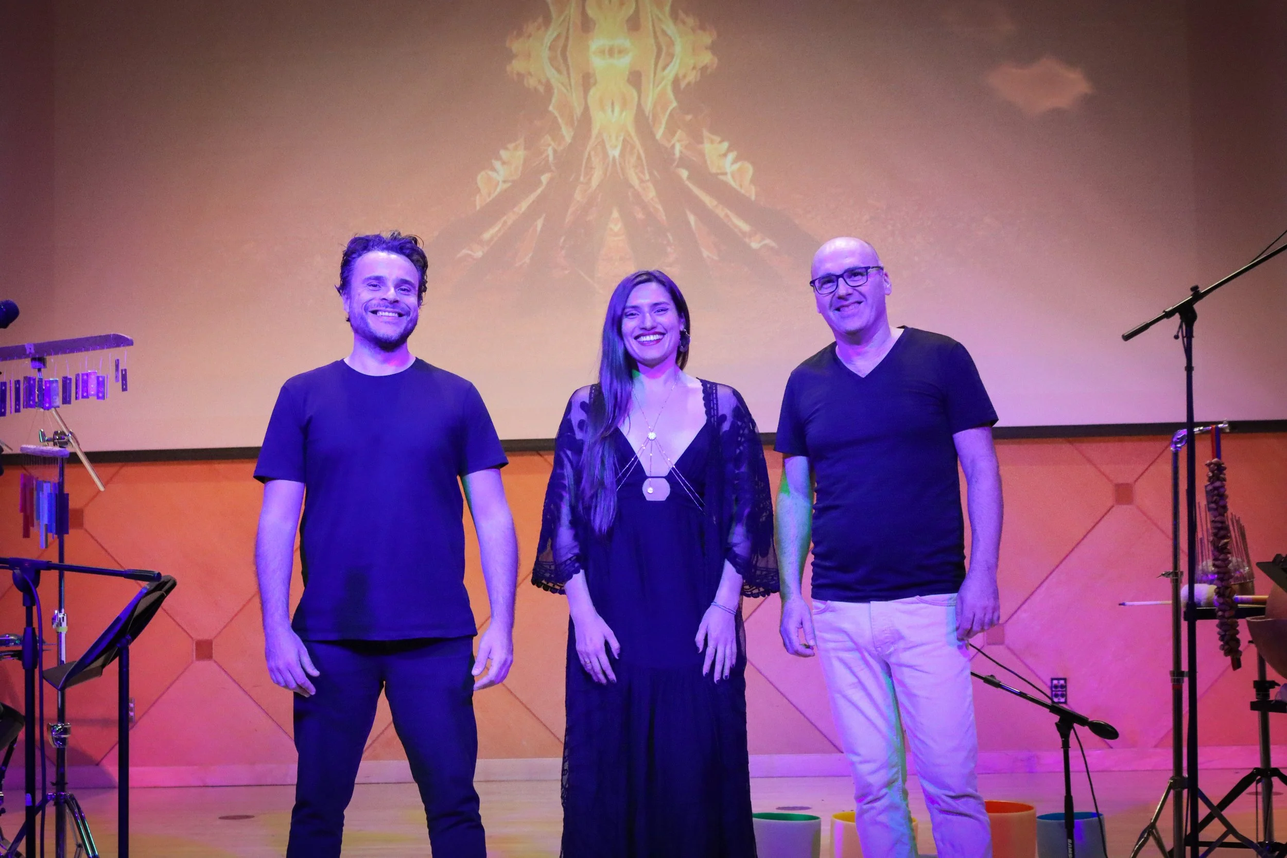 Three people standing on stage, smiling at the camera. A man with dark hair and a beard on the left, a woman with long dark hair and earrings in the middle, and a bald man with glasses on the right. Musical instruments and equipment are visible on st