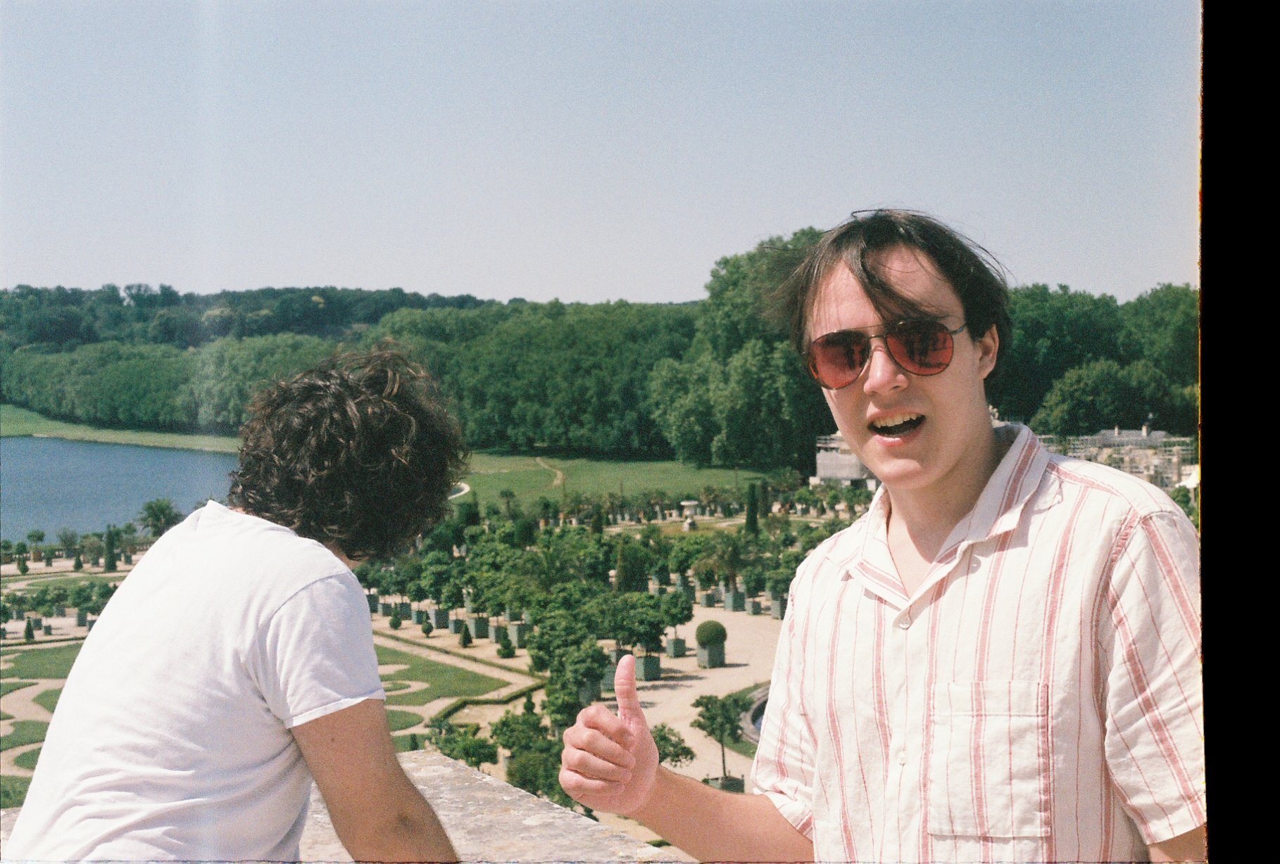 Two young men outdoors during daytime, one facing away and the other giving a thumbs-up, with lush green trees, a lake, and a garden in the background.