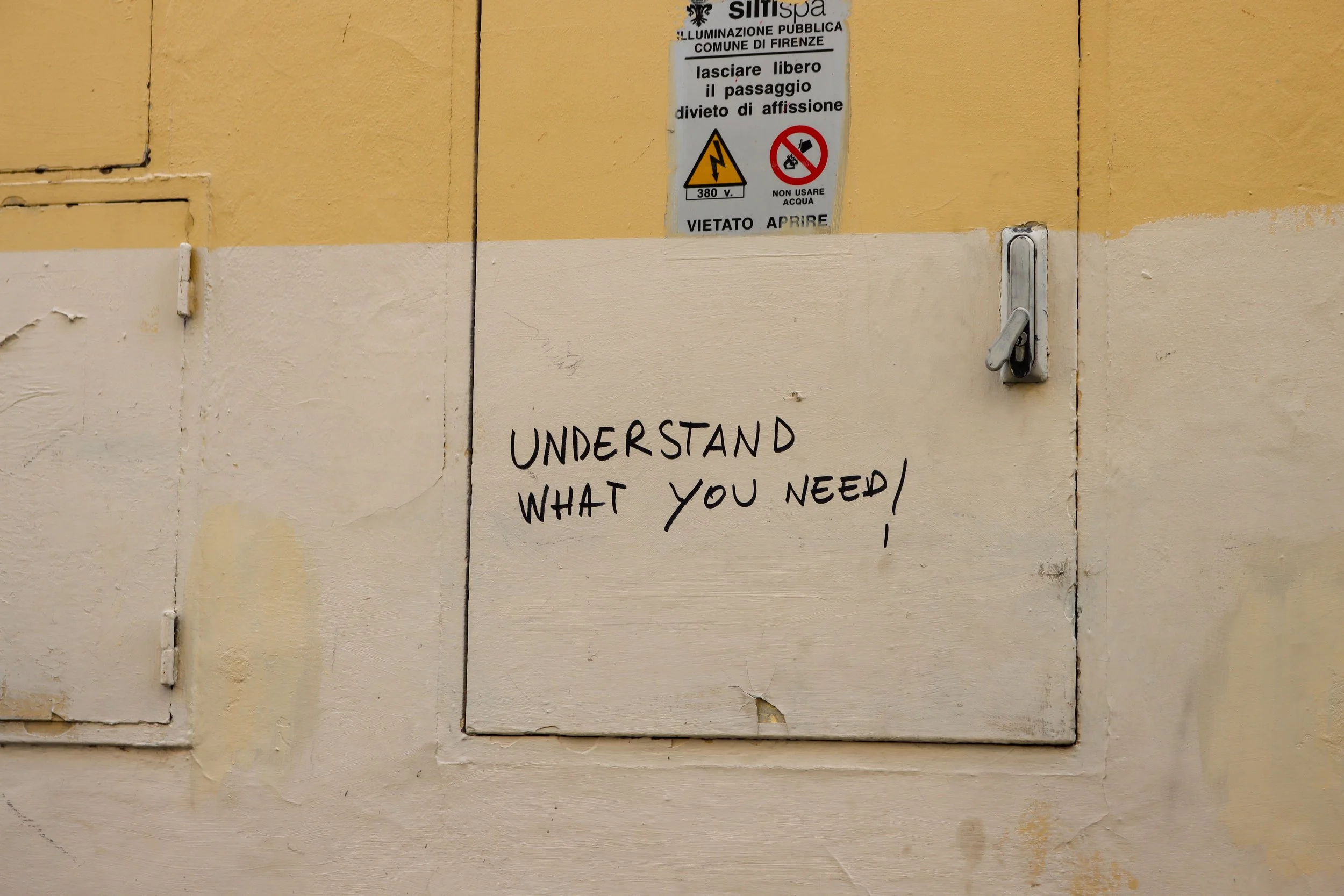 A yellow and cream colored wall with a white electrical panel door. Above it, there is a public lighting sign from Florence, Italy, with multiple warnings including no fireworks and electrical hazard symbols. Below is a handwritten sign that says, "U