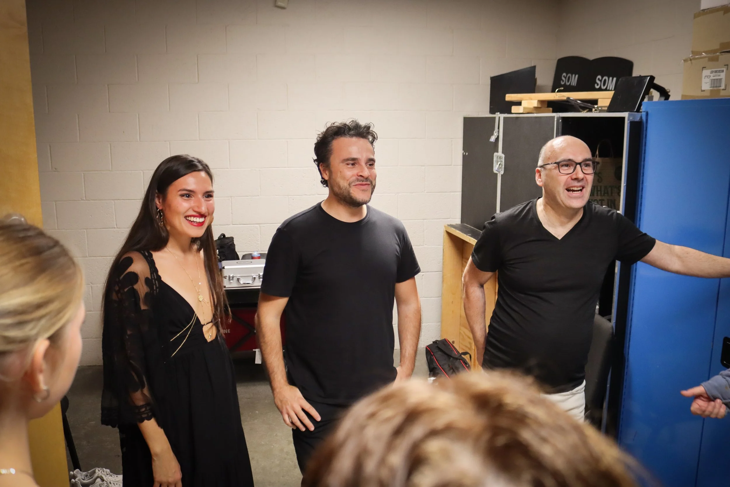 Three adults standing in a room with a brick wall, engaging with children who are partially visible in the foreground. The woman on the left is smiling and wearing a black dress, the man in the middle is smiling and wearing a black t-shirt, and the m