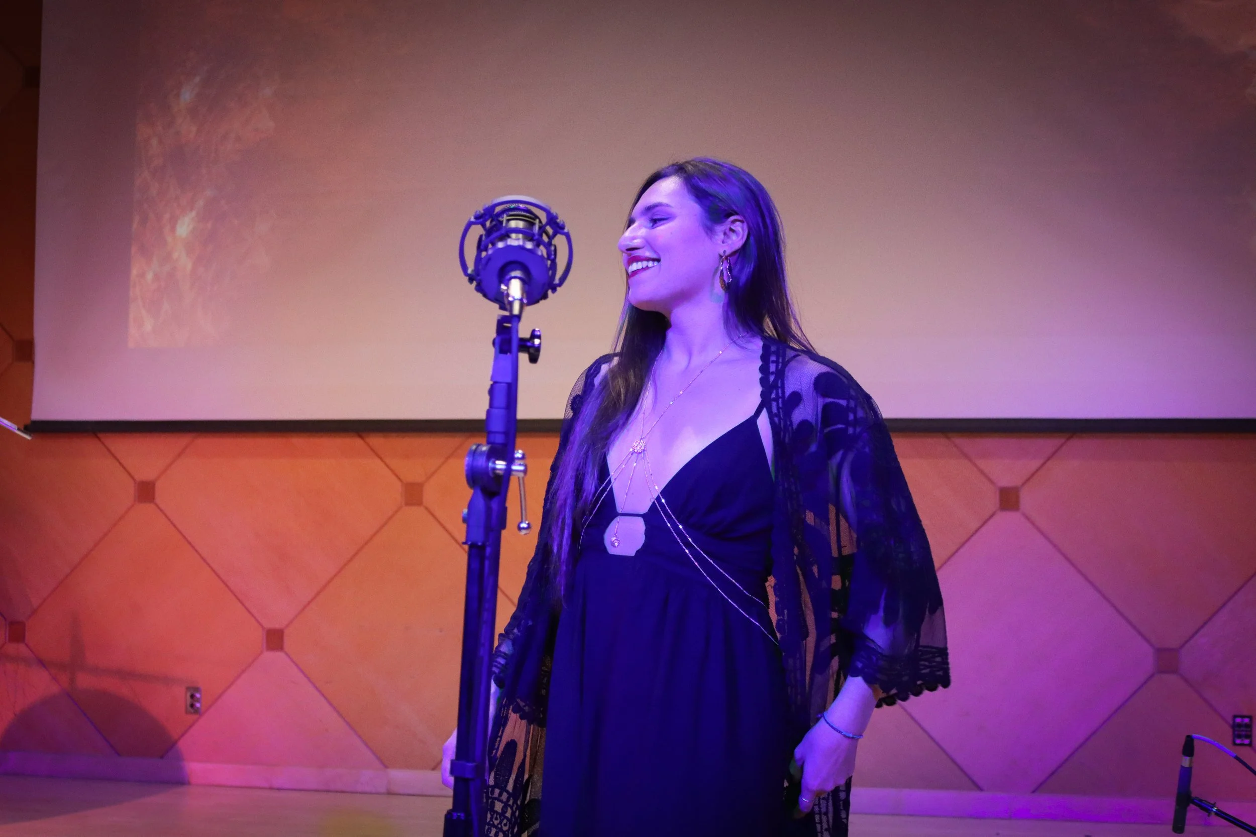 A woman with long hair smiling at a microphone on stage, wearing a black dress with lace sleeves and layered necklaces, with a colorful gradient background.