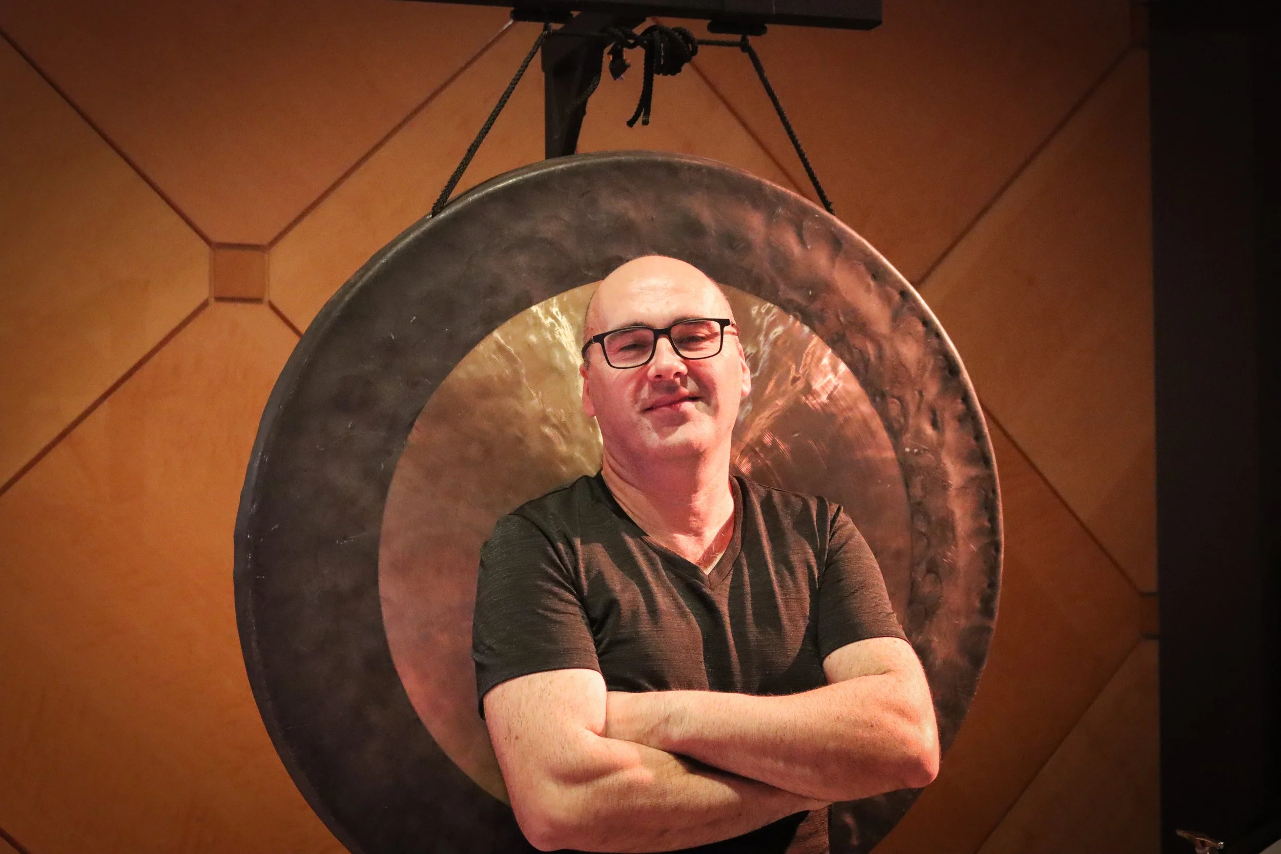 A man with glasses and a black shirt standing with arms crossed inside a large circular metallic frame, in front of a wooden paneled wall.