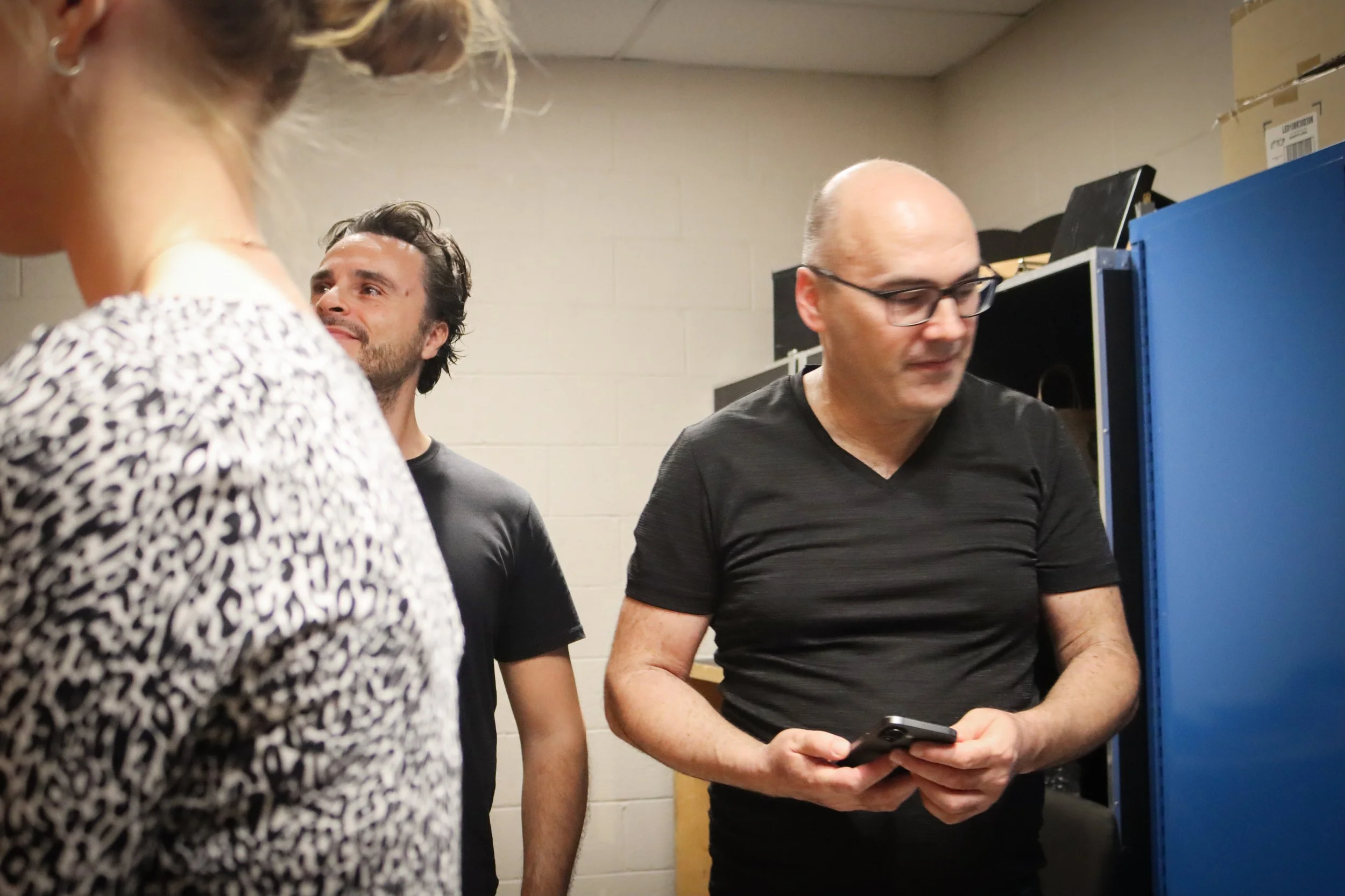 Three people standing in a room with beige brick walls, two men and one woman. One man with glasses is looking at his phone, another man with darker hair and beard is looking at someone or something outside the frame, and the woman with earrings has 
