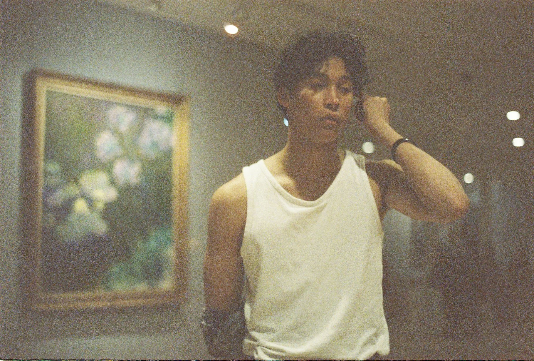 A woman with dark, curly hair wearing a white sleeveless top, standing indoors with her hand near her ear, in front of a wall with a framed floral painting, and ceiling lights visible.