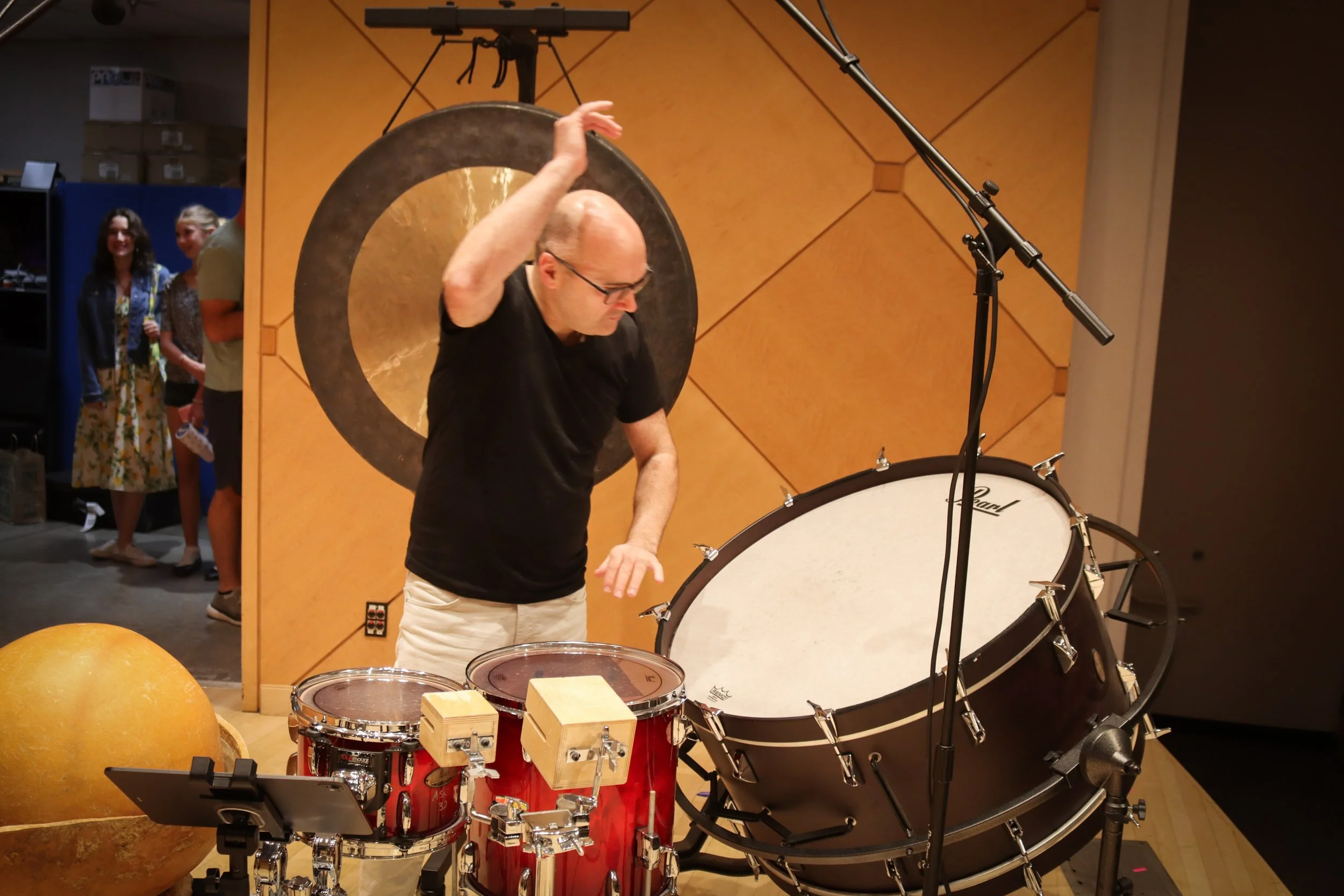 A man with glasses and a black shirt playing percussion instruments, including a drum and a large gong, in a recording studio or music performance space.