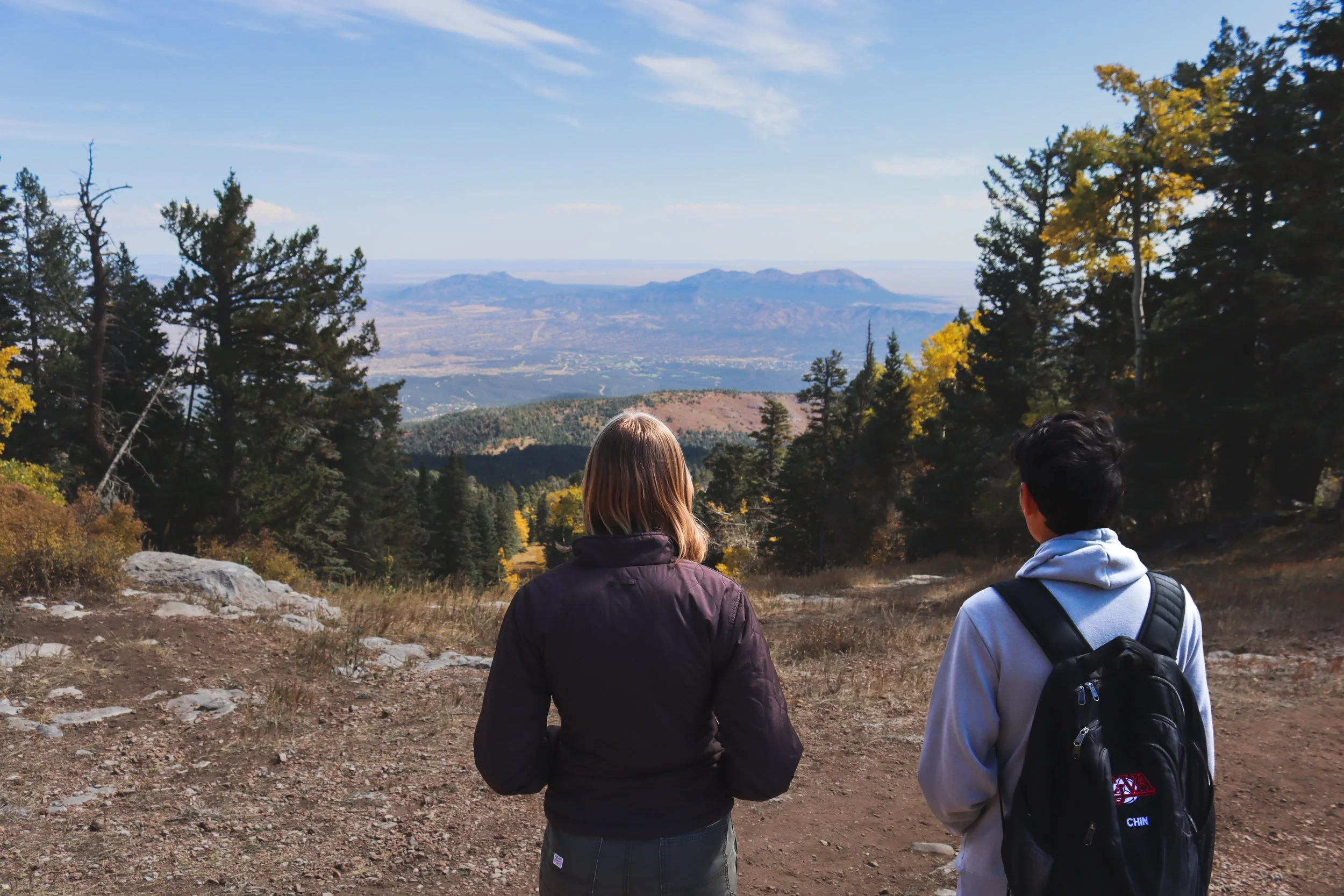 Two people standing outdoors on a hiking trail, looking at a scenic mountainous landscape with trees and a distant mountain range under a blue sky.
