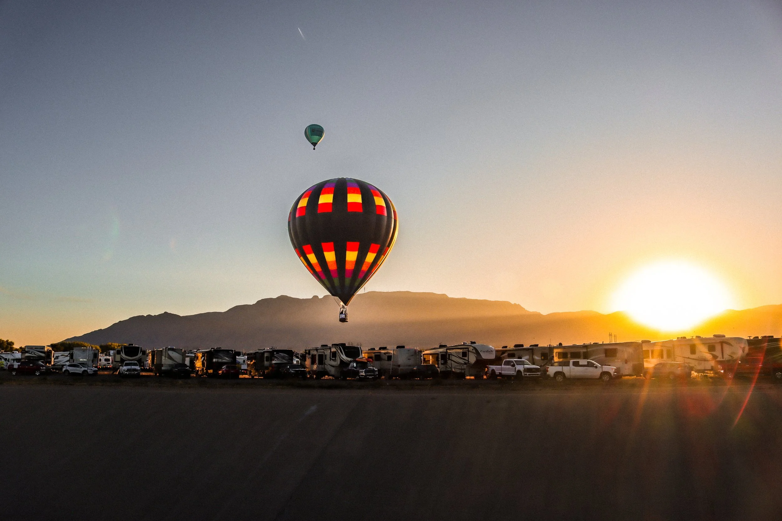 Hot air balloons floating in the sky at sunset over a landscape with mountains and a parking lot filled with RVs and cars.