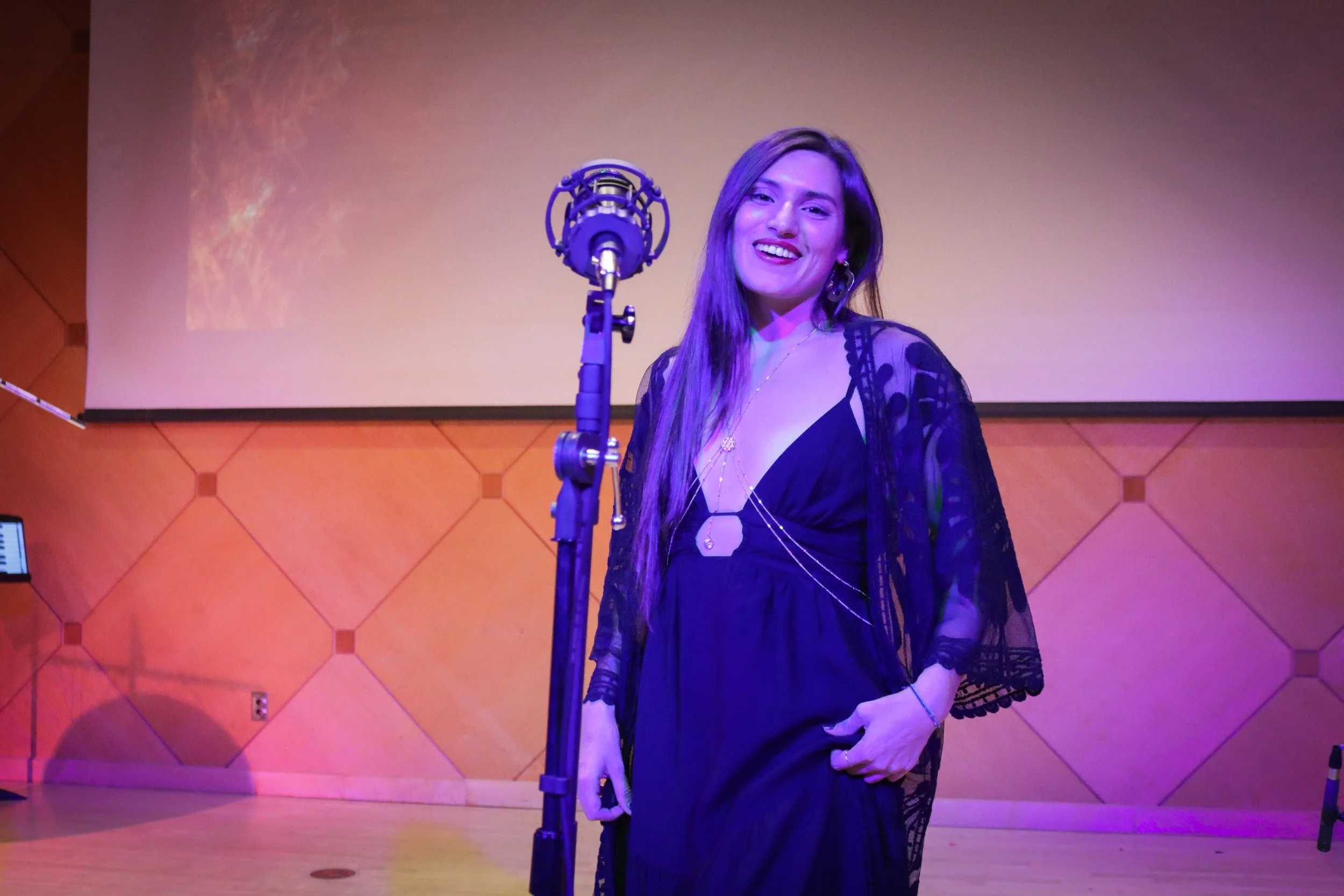 A woman with long brown hair smiling on stage in front of a microphone, wearing a black dress with lace sleeves, in a room with wood-paneled walls and a large blank projection screen.