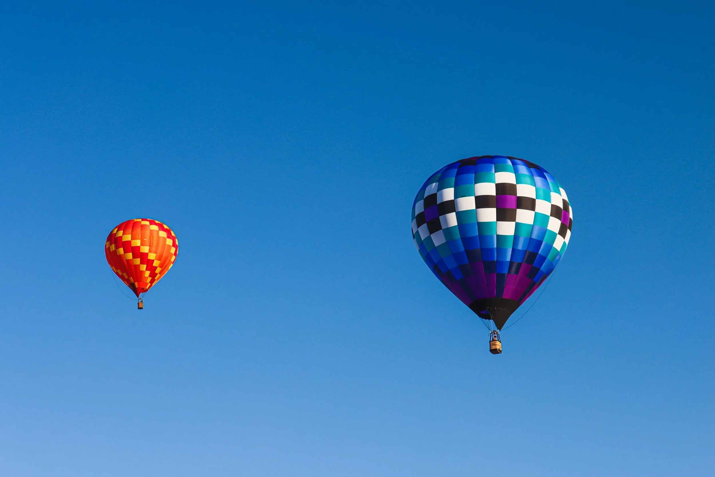 Two colorful hot air balloons flying in a clear blue sky, one with a red and yellow checkered pattern and the other with a blue, black, white, and purple checkered pattern.