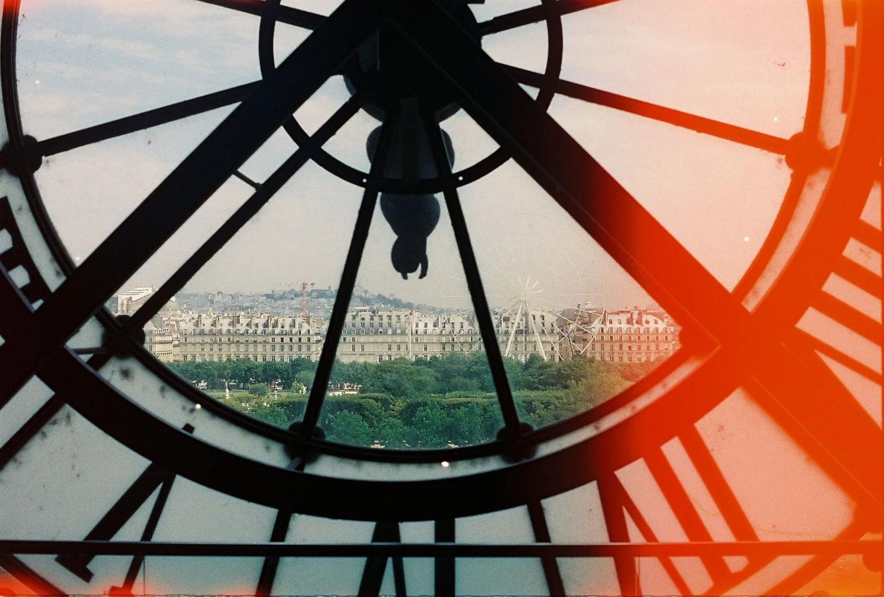 View of a cityscape with buildings, trees, and a Ferris wheel, seen through the clock face of a large, ornate clock tower from inside.