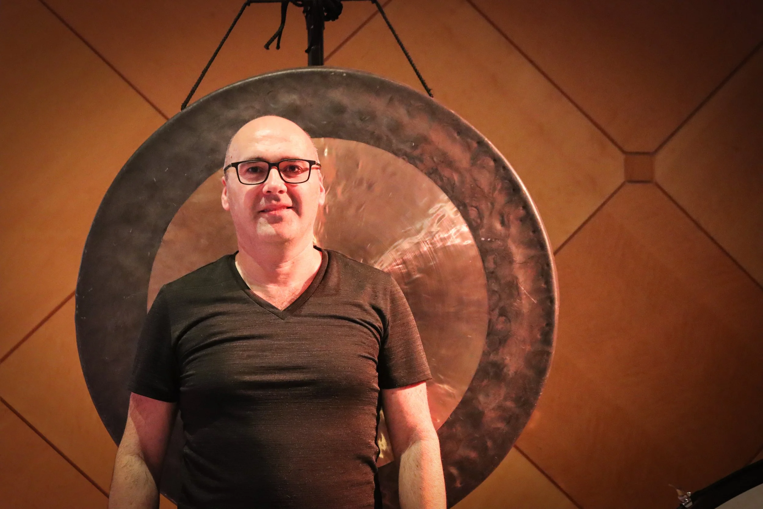 A man with glasses and a bald head standing in front of a large metallic gong with a wooden wall background.