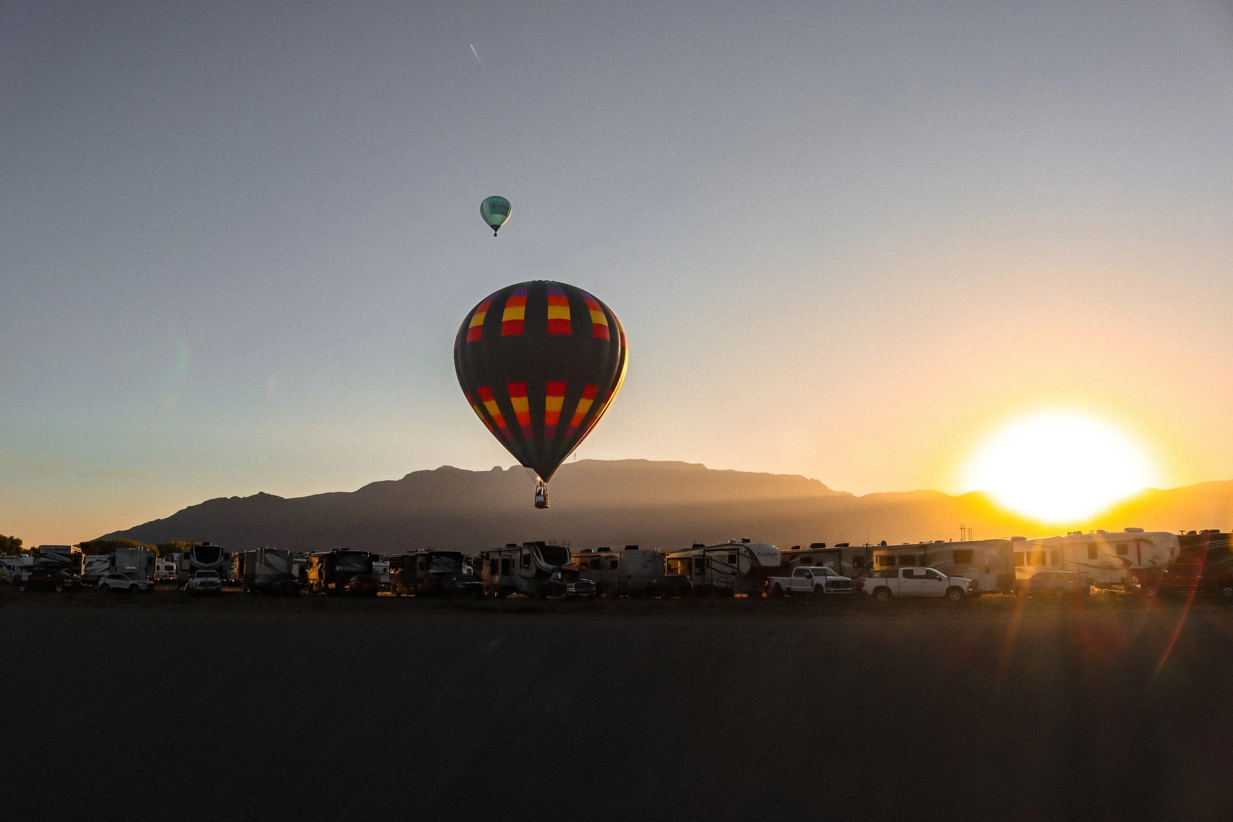 Two hot air balloons floating in the sky during sunset over a landscape with mountains. The balloon in the foreground has a colorful checkered pattern, while the balloon in the background is primarily blue.