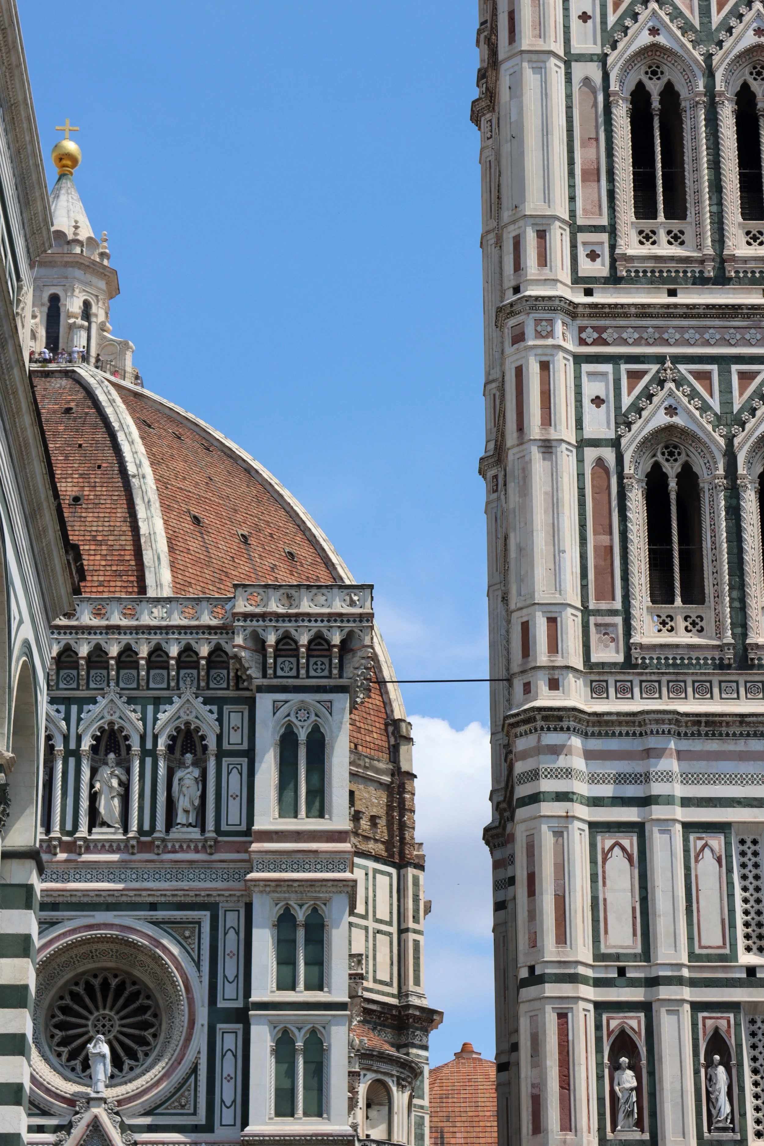 Close-up view of the intricate marble facade of Florence Cathedral's Cathedral of Santa Maria del Fiore, including detailed carvings, statues, and colorful stone patterns, with part of the red-tiled dome visible on the left side of the image.