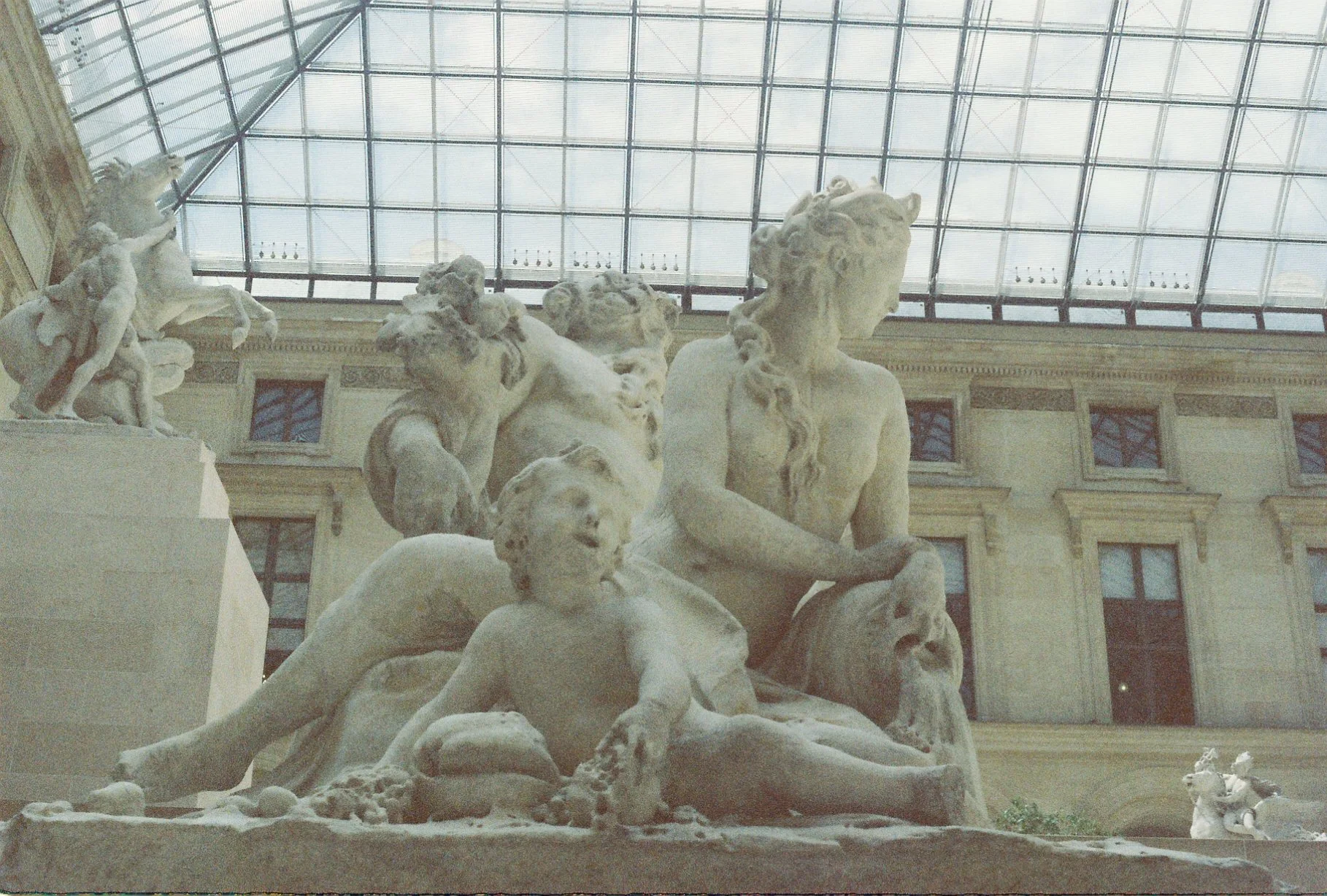 Museum sculpture of mythological figures including a seated woman with a lion's head, a lion, and other figures, beneath a large glass ceiling with metal framework.