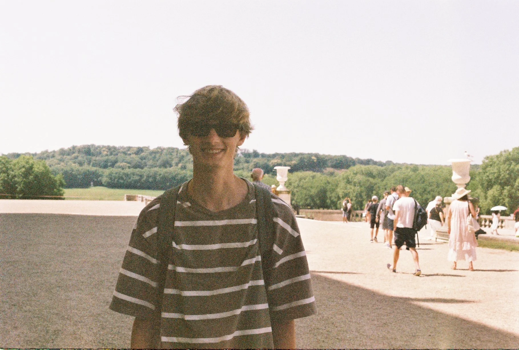 A person with short hair, wearing sunglasses, a striped shirt, and a backpack, standing outdoors with a large group of people in the background, some walking and others holding umbrellas, near a scenic area with green trees and a stone balustrade.