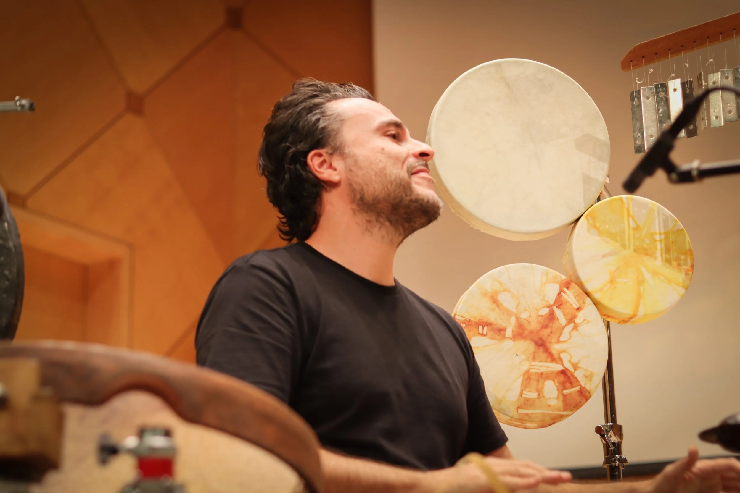 A man with dark hair and a beard playing drums in a music studio, with decorative circular panels in the background.