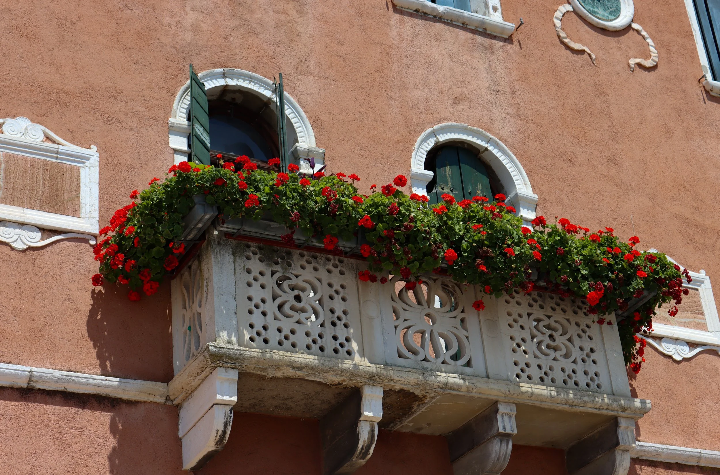 A peach-colored building with two small arched windows and a decorative white balcony with red flowers, and green shutters on the windows.