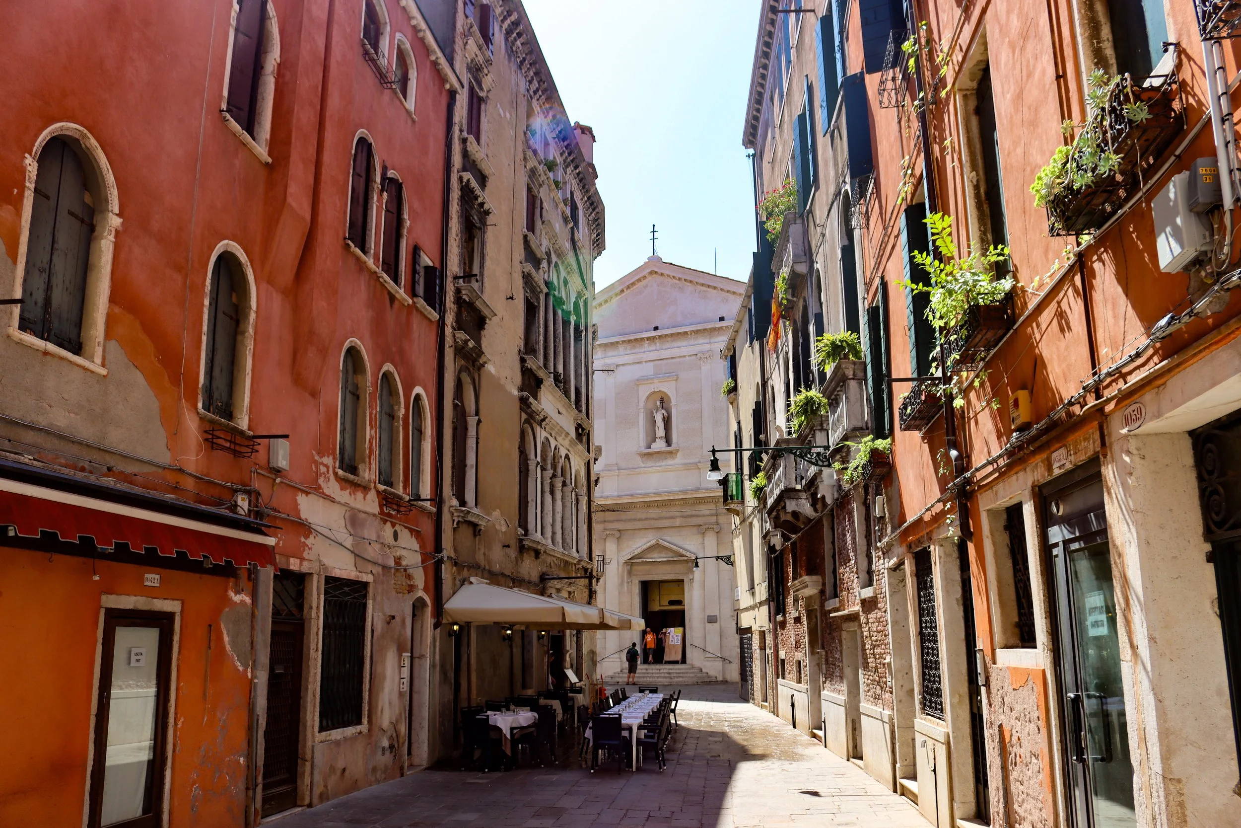 A narrow European street with colorful old buildings on both sides, some with plants on balconies, leading to a church or historic building with a cross on top, under a clear sunny sky.