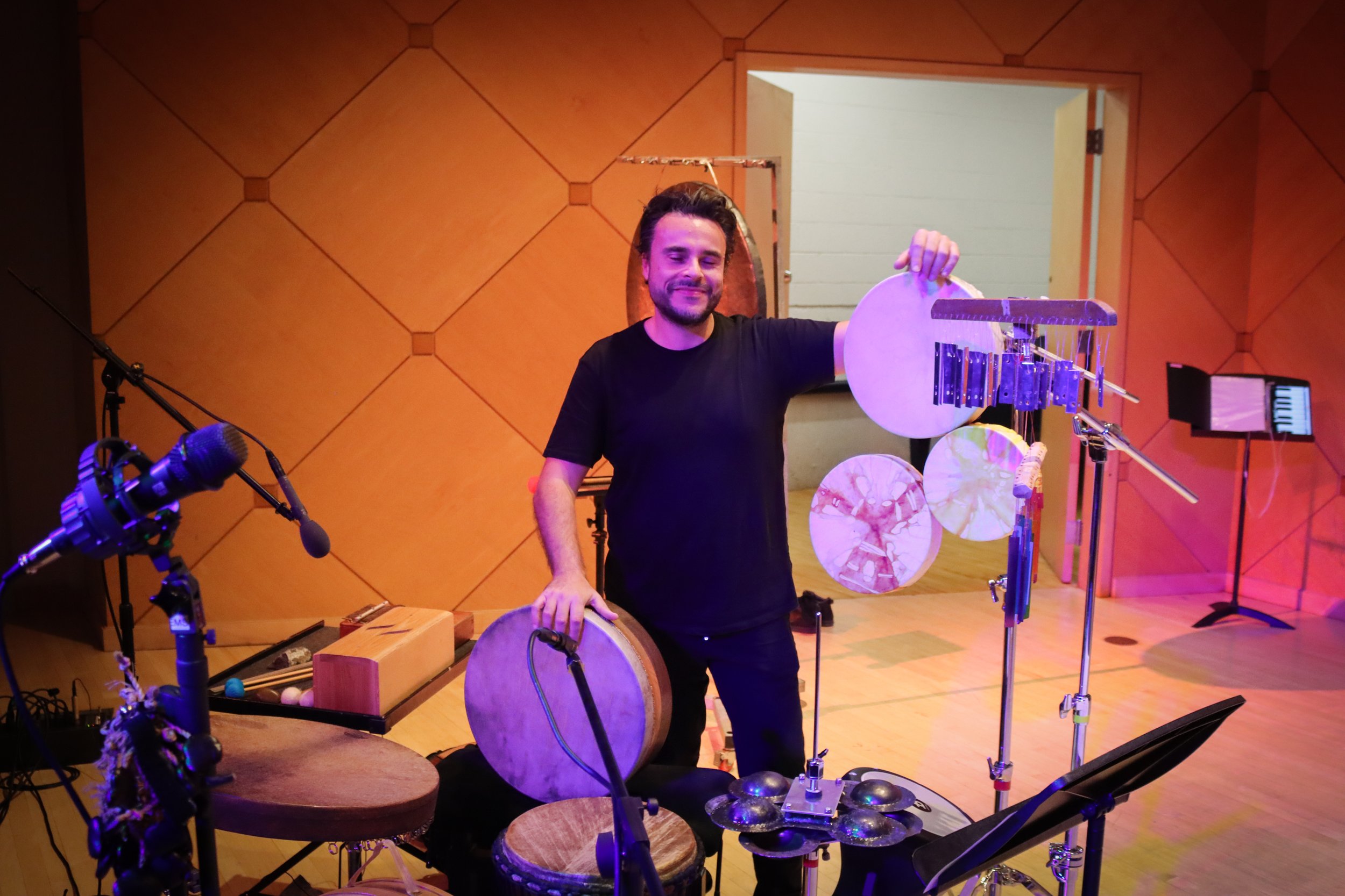 A man with dark hair and a beard smiling and holding a percussion instrument in a music studio or performance space, surrounded by drums, microphones, and other musical equipment with wooden acoustic panels on the wall.