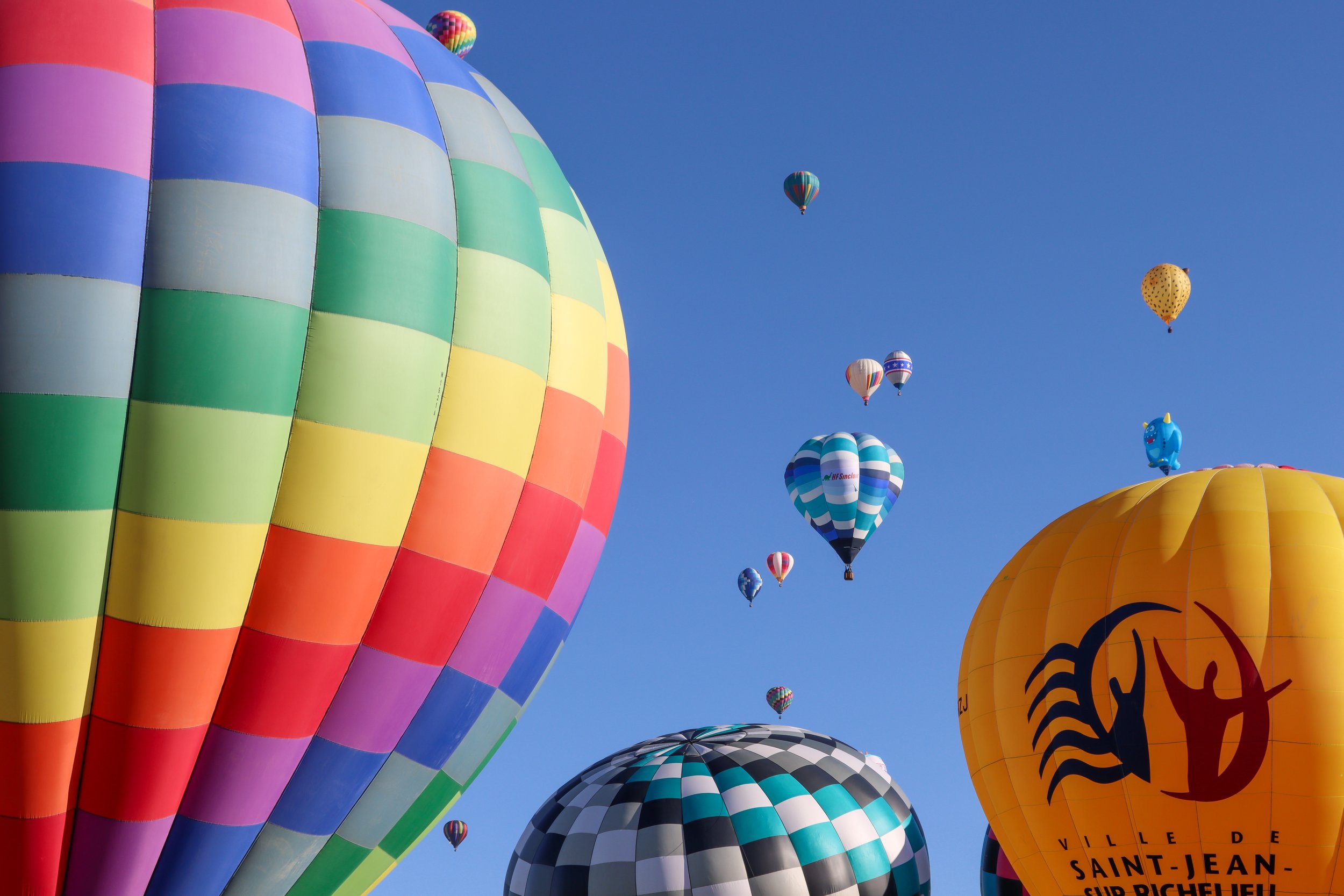Multiple colorful hot air balloons flying in a clear blue sky, including a rainbow-patterned balloon, a yellow balloon with tiger stripes, and other patterned balloons.
