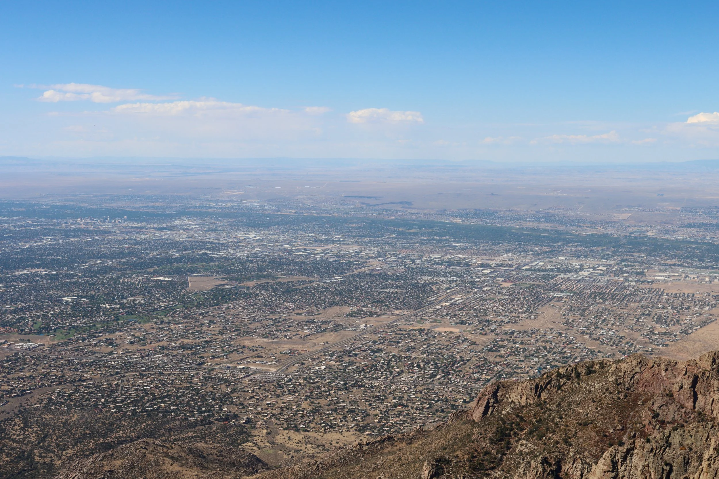 Aerial view of a cityscape with a sprawling urban area, fields, and mountains in the distance, taken from a high vantage point on a clear day.