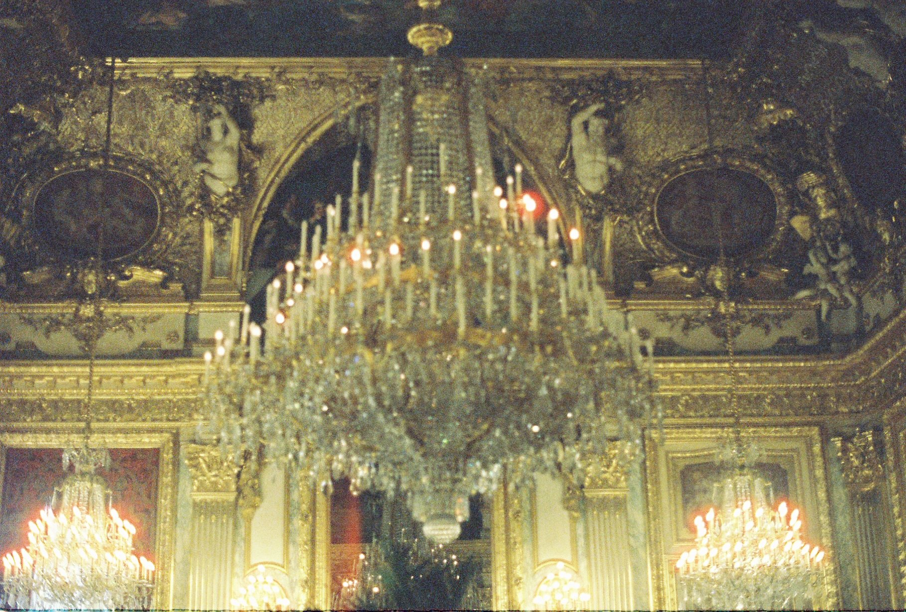 Elegant interior of a grand room with ornate gold detailing, a large chandelier hanging from the ceiling, and wall portraits.