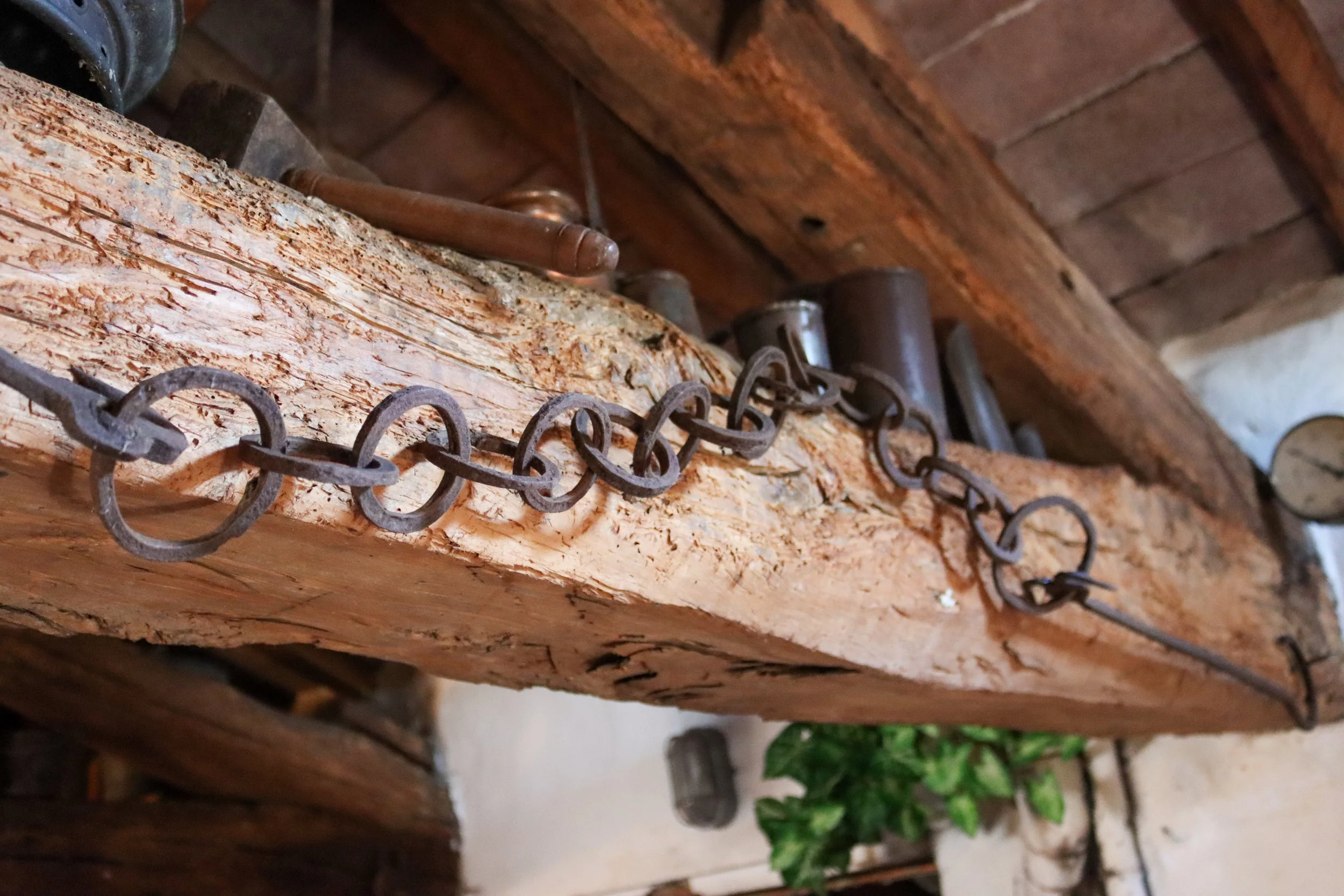 Close-up of a rusty chain attached to a piece of wood with a wooden handle and a metal pipe, in front of a wooden ceiling and a brick wall.