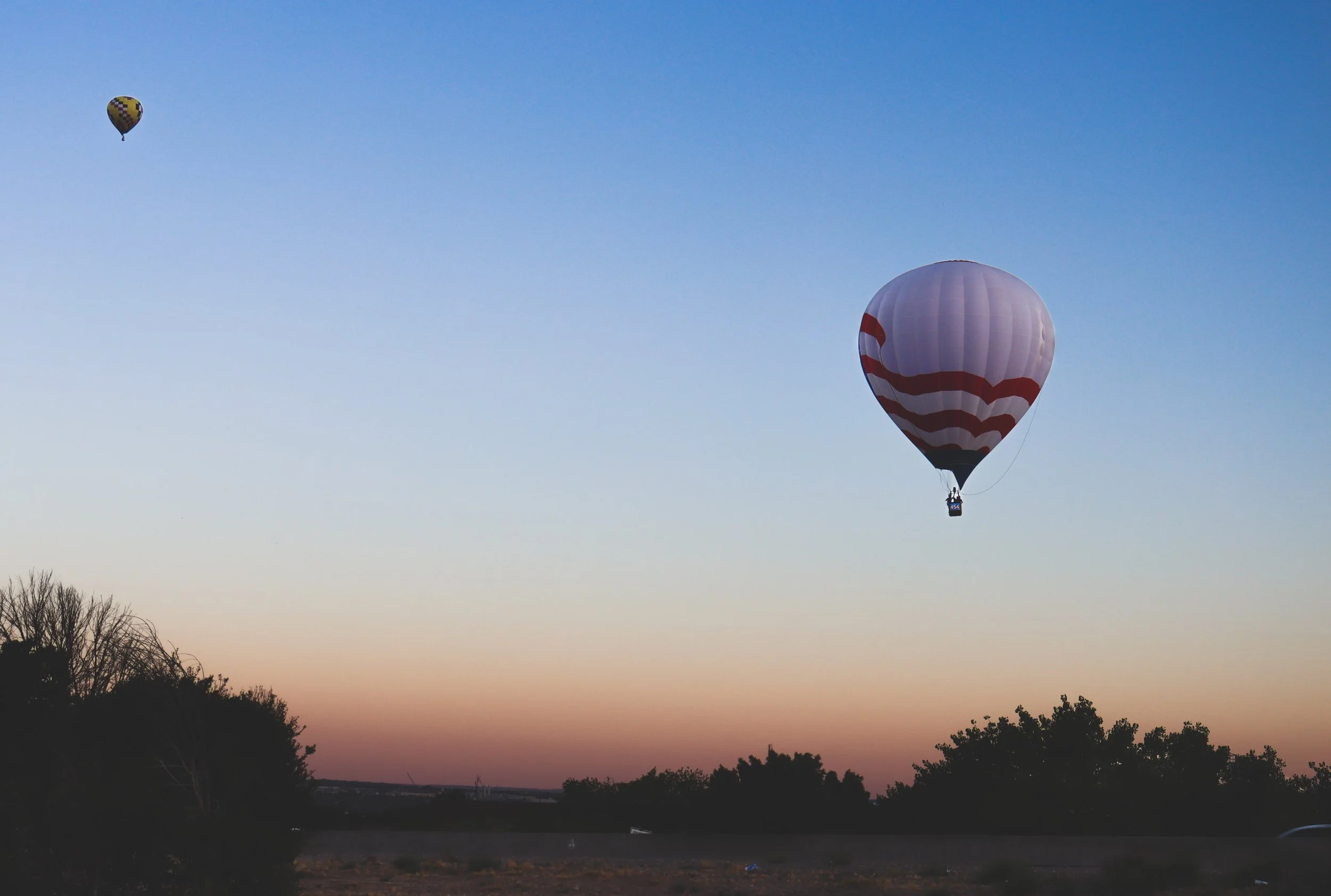Two hot air balloons floating in the sky during sunset, with one balloon mostly pink with red and white stripes, and the other smaller and primarily yellow with a checkered pattern, above a silhouette of trees and fields.