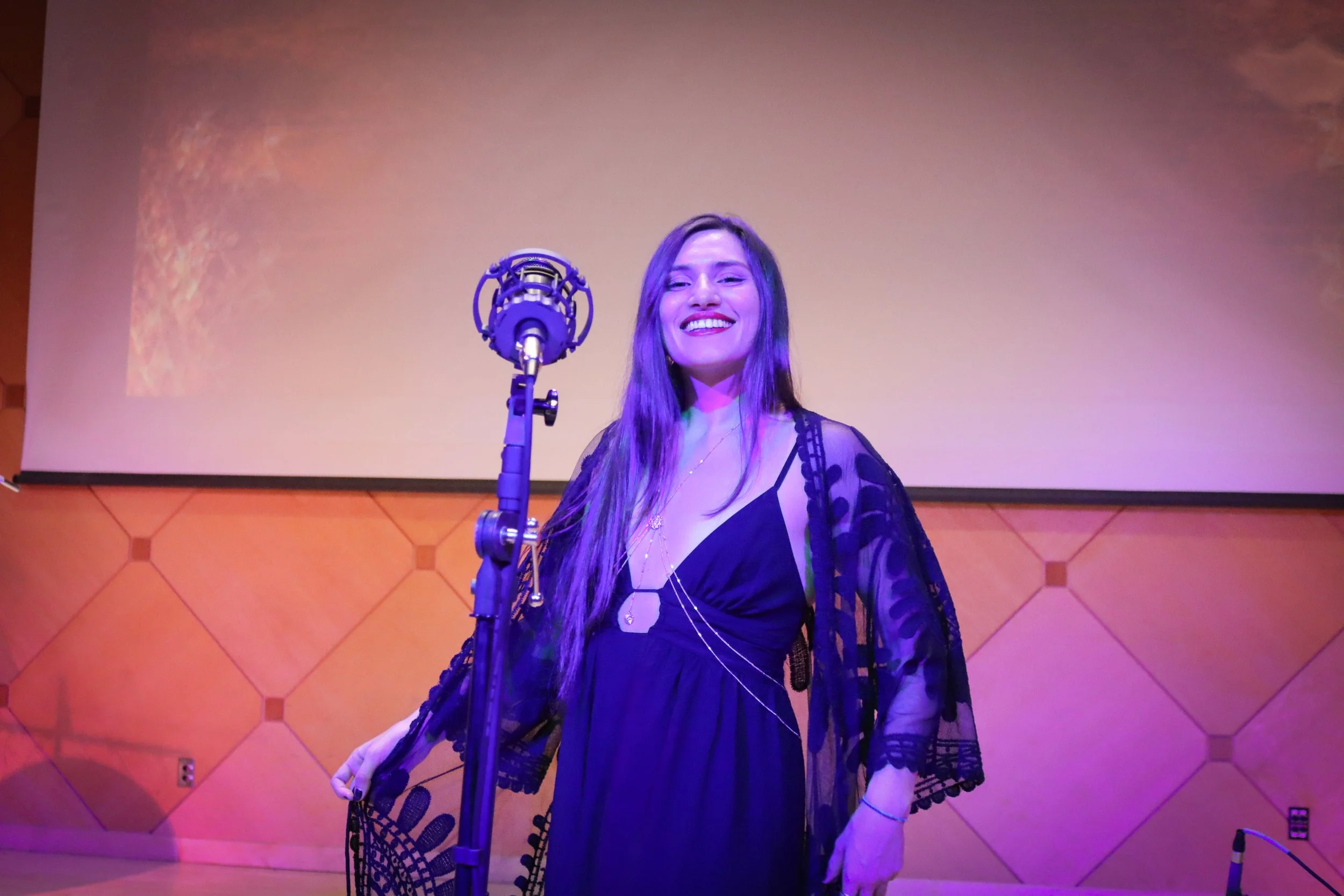 A woman with long brown hair, smiling, standing in front of a microphone on a stage with a wooden-tile wall background lighting in purple and pink tones.
