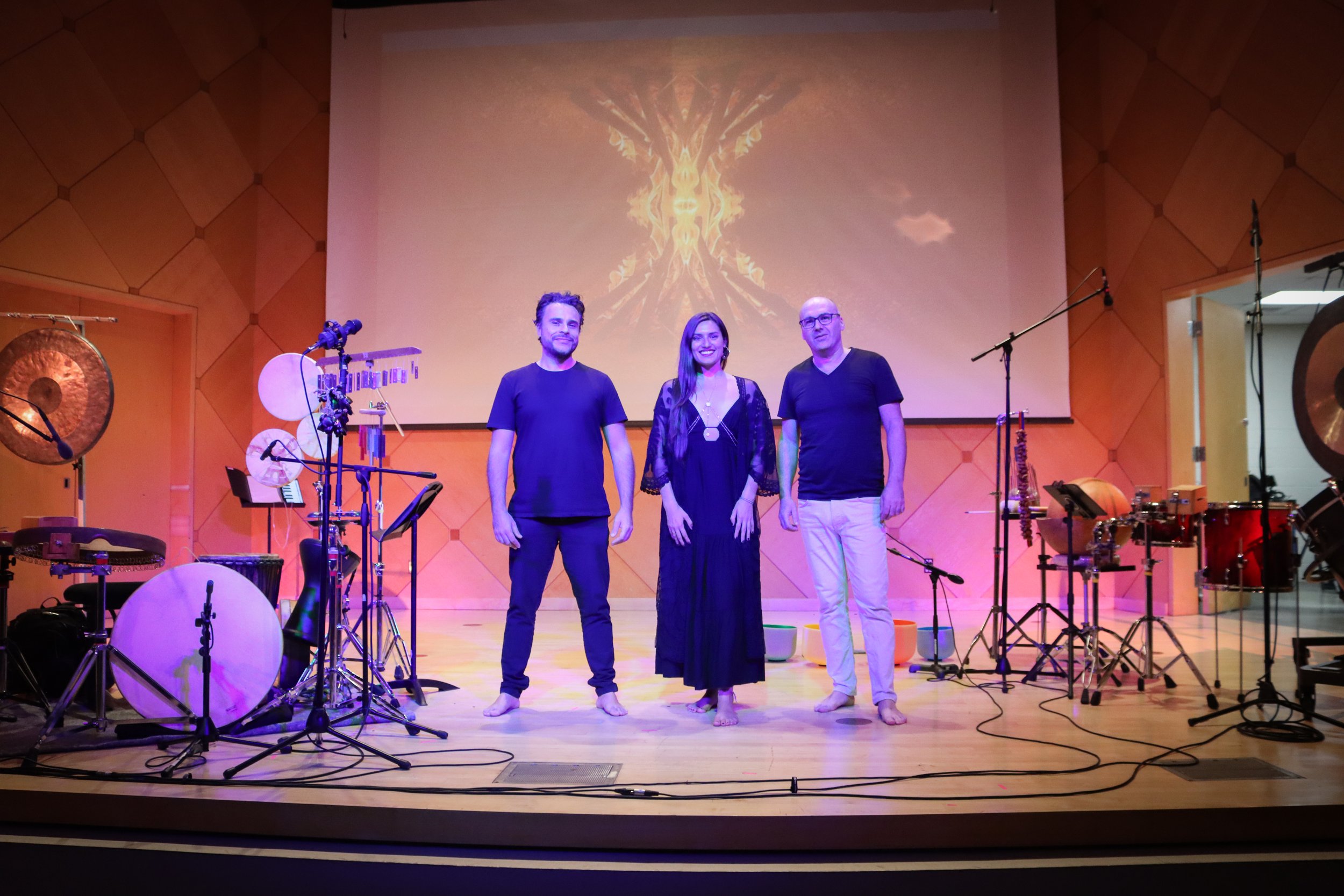 Three performers standing barefoot on a stage with musical instruments around them, in front of a large screen displaying abstract artwork, with drums and other percussion instruments visible.