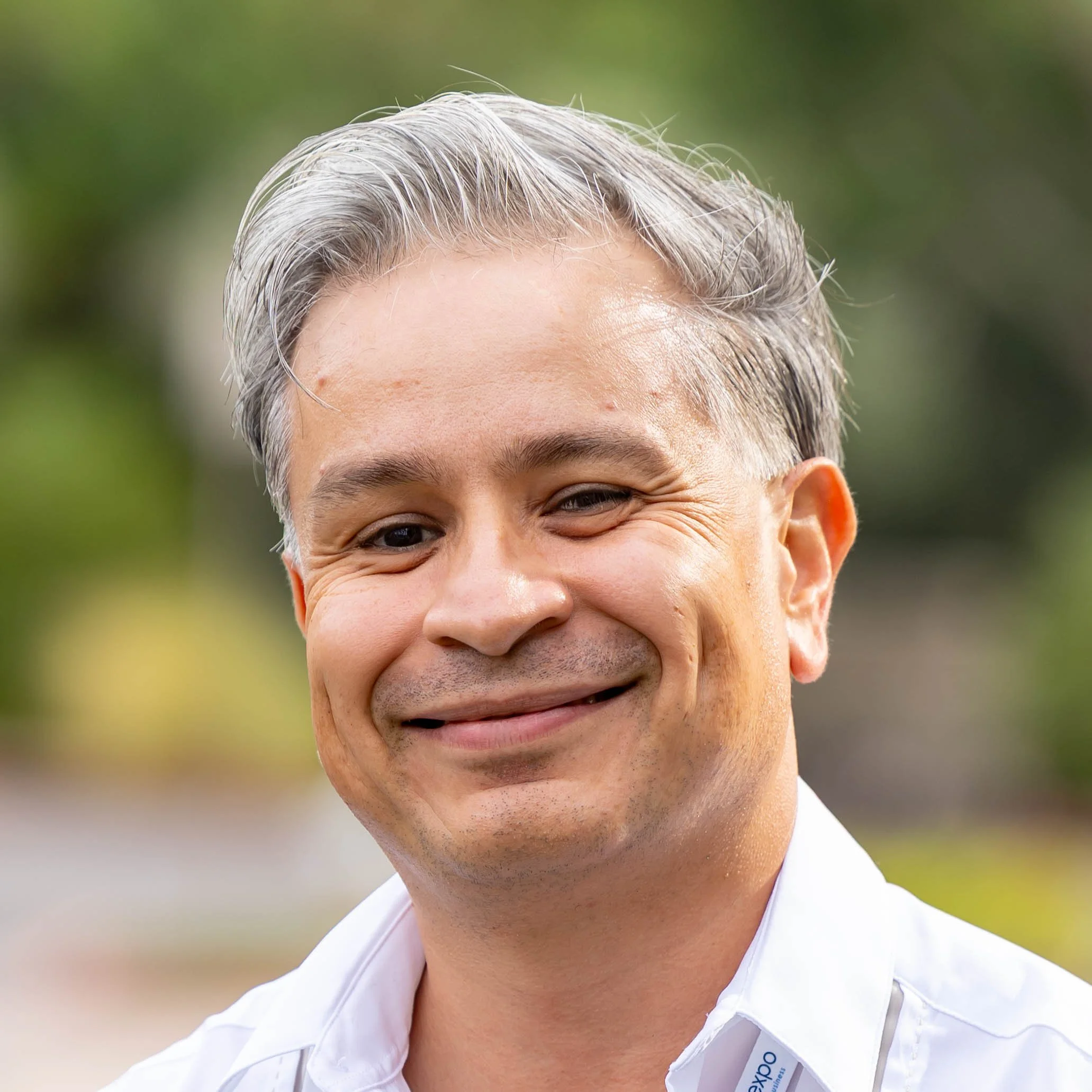 Close-up of a smiling man with short gray hair, wearing a white shirt, with a blurred green background.