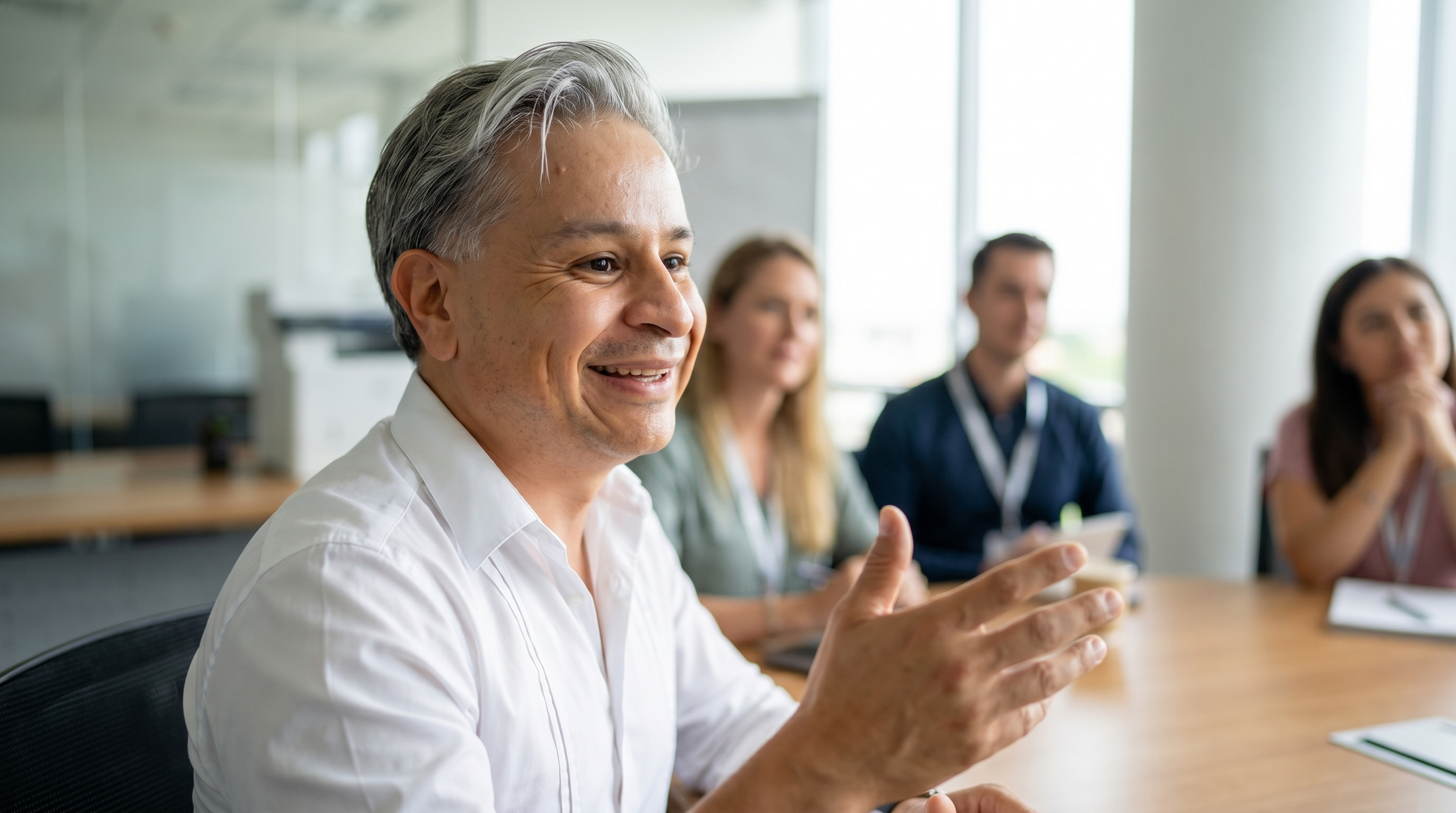 A group of diverse people in a meeting room, with a smiling man in focus speaking or presenting.