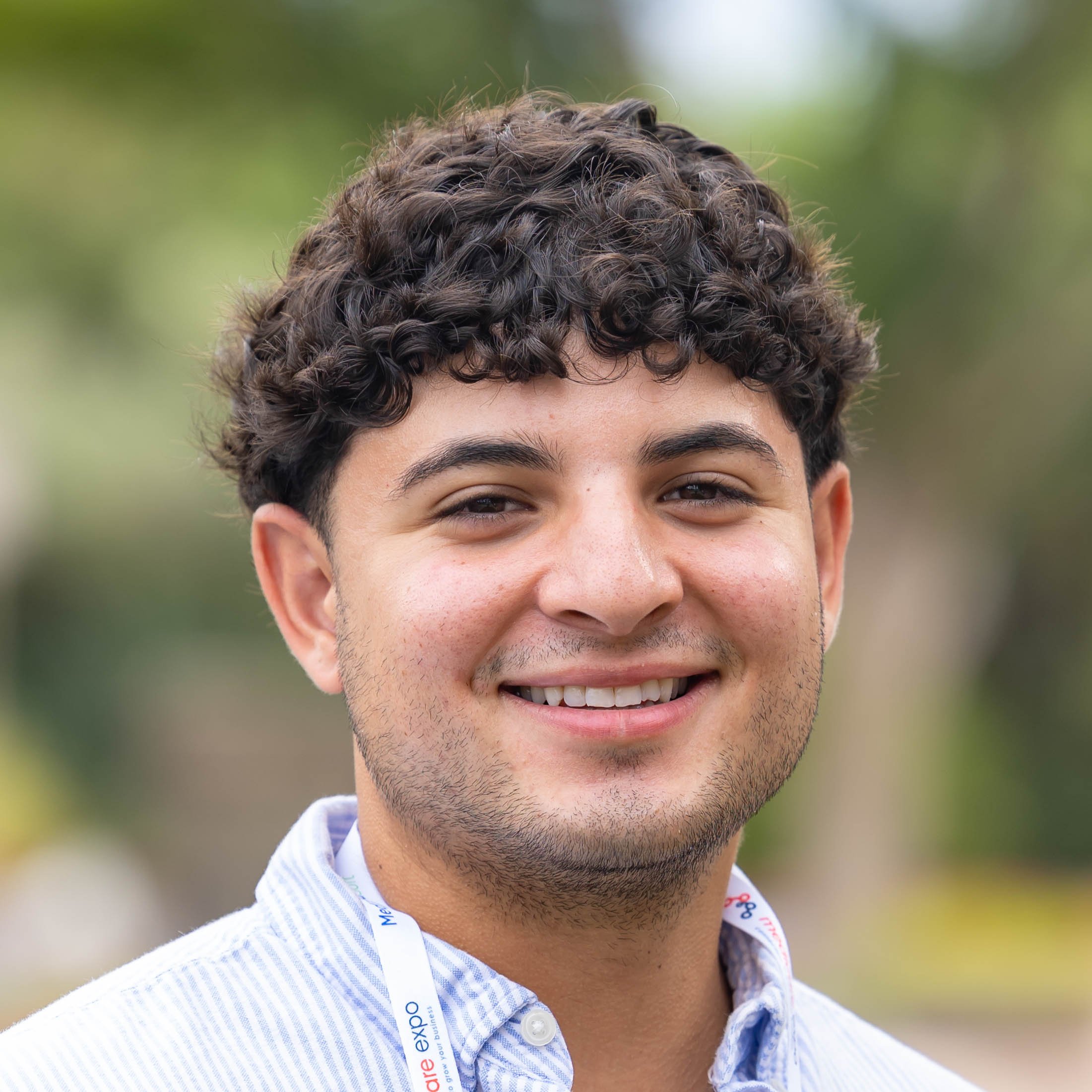 Close-up of a young man with curly dark hair, brown eyes, and light facial hair, smiling outdoors with a blurred green background.