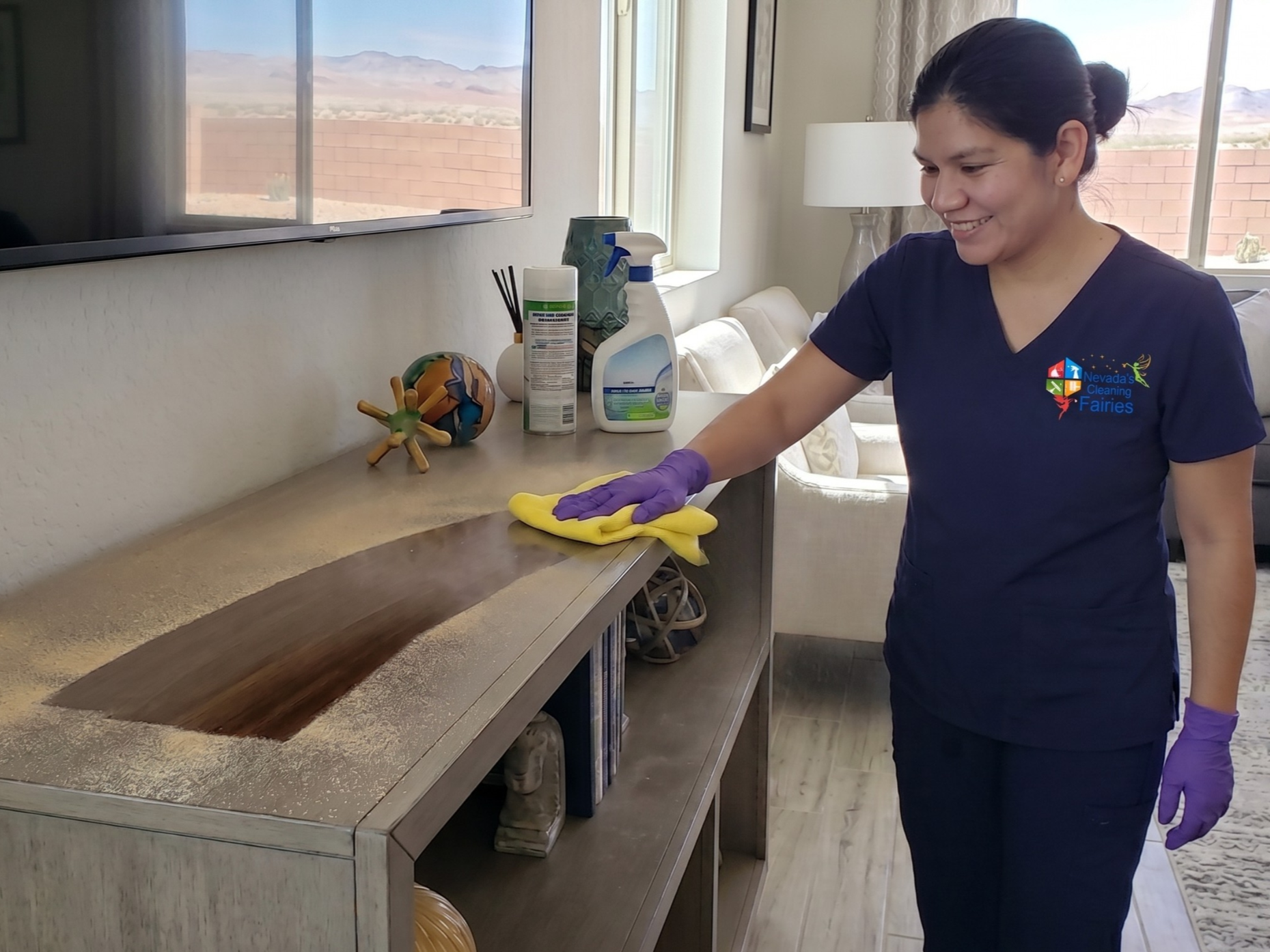 A smiling cleaner in a Nevada's Cleaning Fairies uniform wipes a thick layer of dust from a dark wood table with a yellow cloth, revealing a clean, shiny surface underneath.