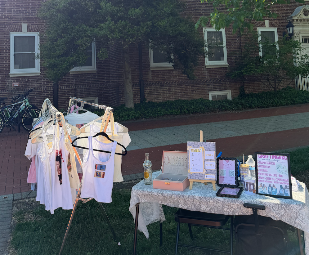 A yard sale setup with a clothing rack holding various T-shirts, and a table with jewelry, signs, and other items on a sidewalk in front of a brick building with trees.