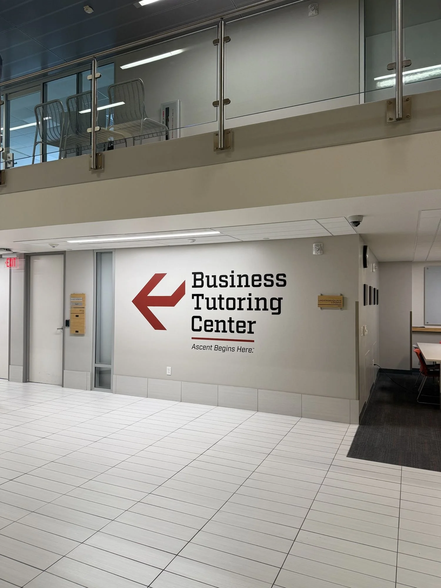School corridor with white tiled floor, a wall with signs pointing to the Business Tutoring Center and undergraduate lounge, and a glassed upper floor with chairs and a security camera.