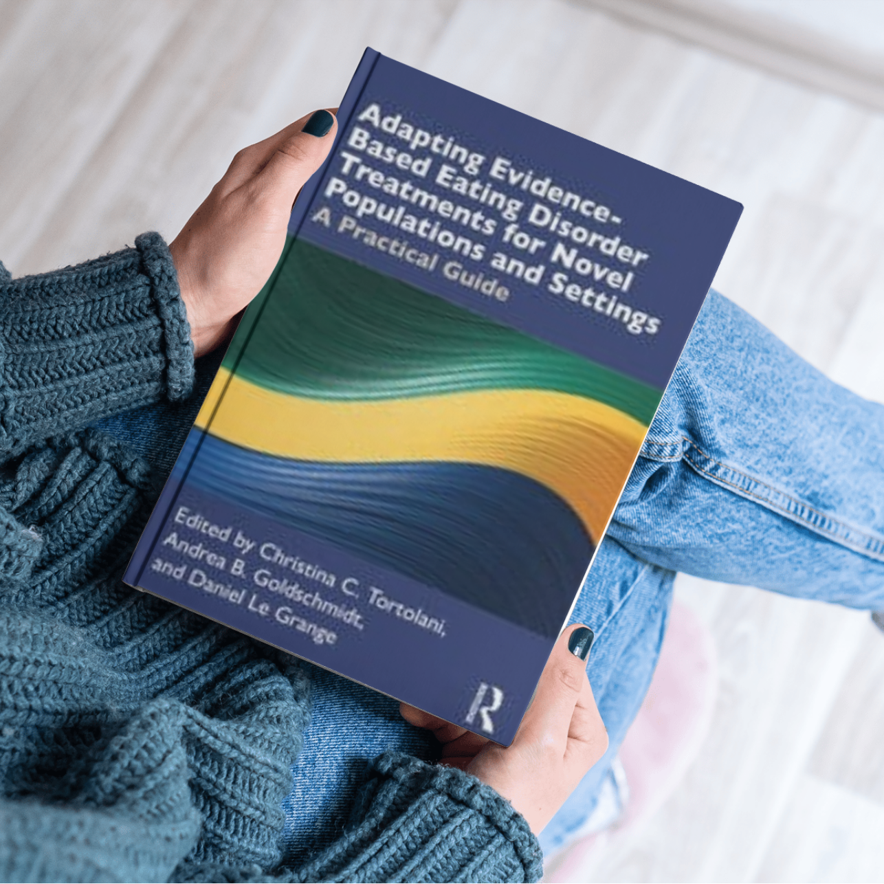 Person wearing a blue knitted sweater and jeans sitting on light-colored wooden floor, holding a book titled 'Adapting Evidence-Based Eating Disorder Treatments for Novel Populations and Settings: A Practical Guide'