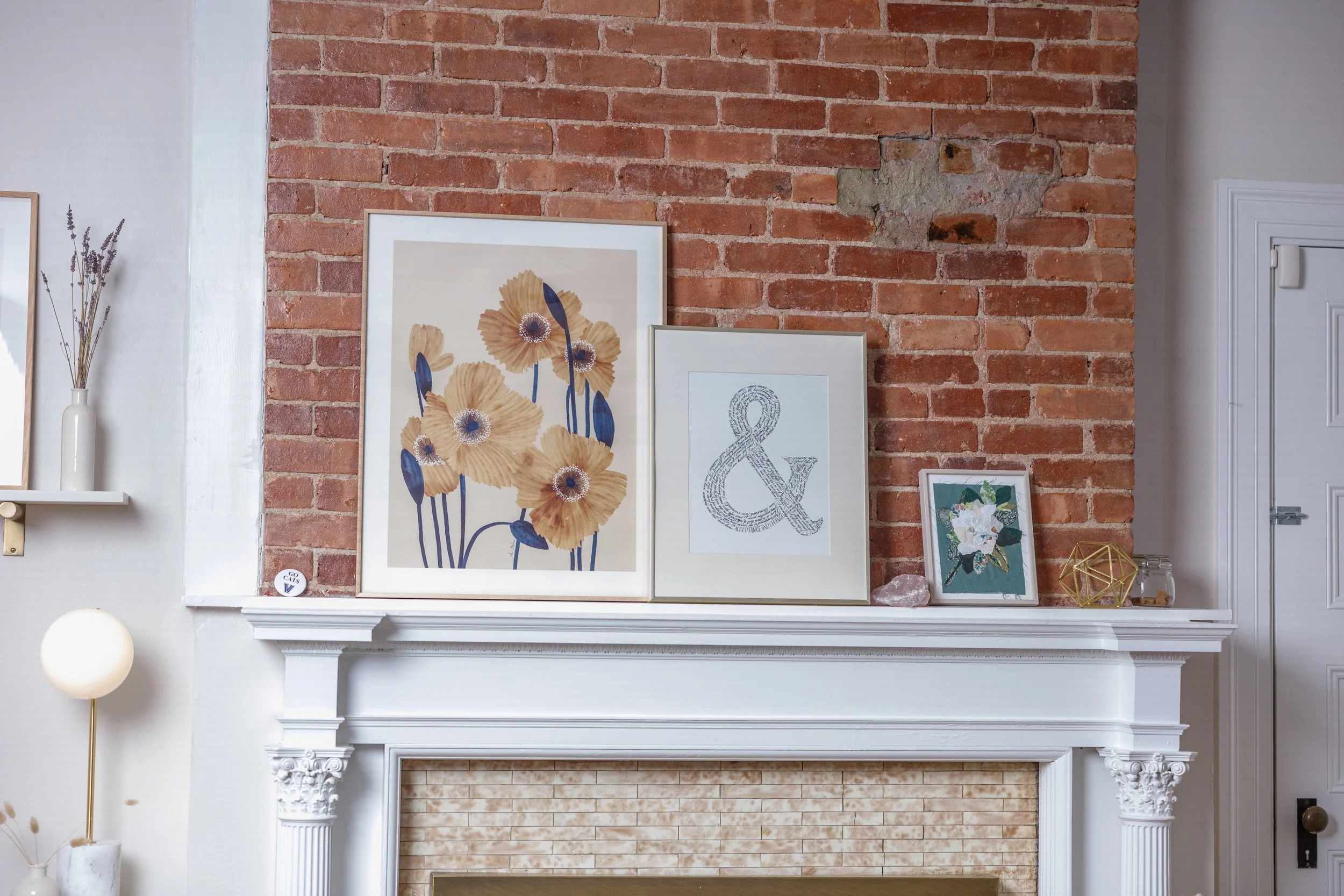 Decorative display on a white fireplace mantel with framed artwork, a small plant, a crystal, and a geometric sculpture against a brick wall background.