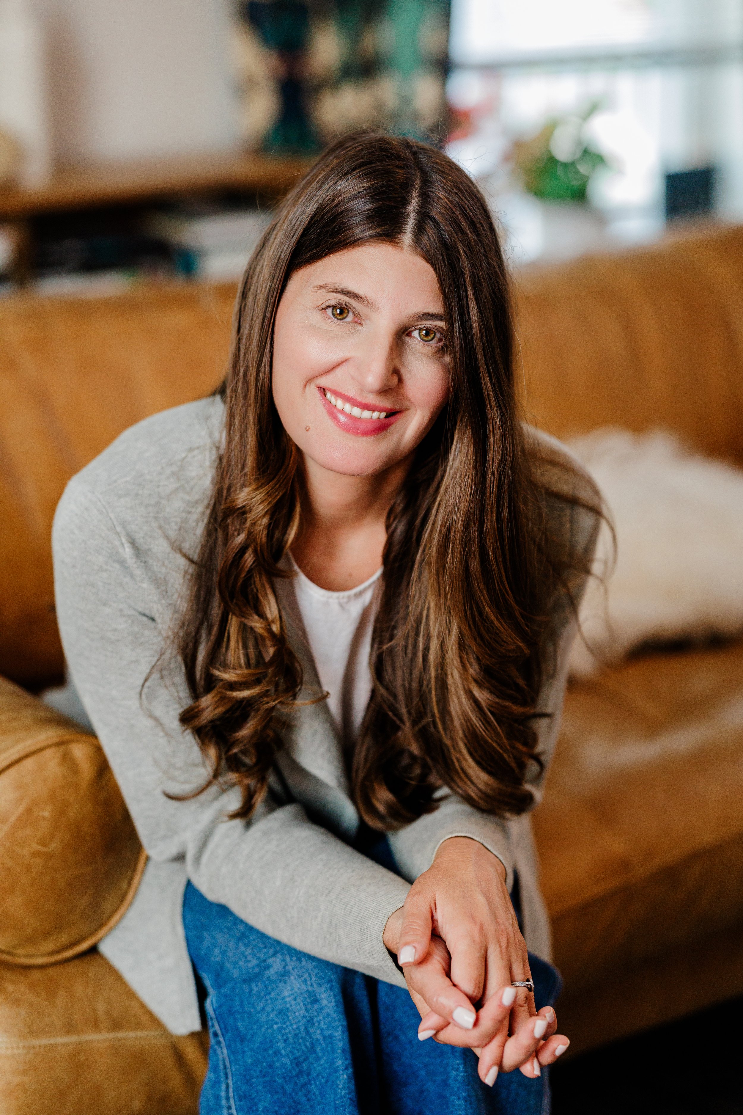 A woman with long brown hair and light skin sitting on a tan sofa, smiling, wearing a light gray hoodie and blue jeans, with a blurred background of a bright room with large windows and various decor.