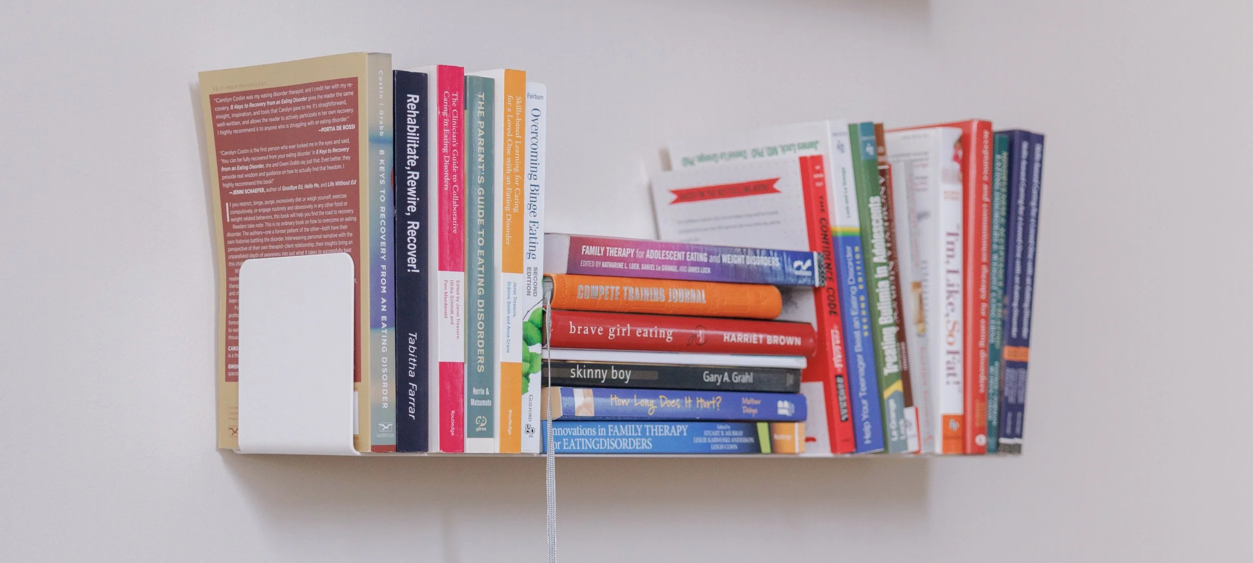 A bookshelf filled with medical and psychology books, arranged vertically, with a small stack of books on top of others, set against a plain white wall.