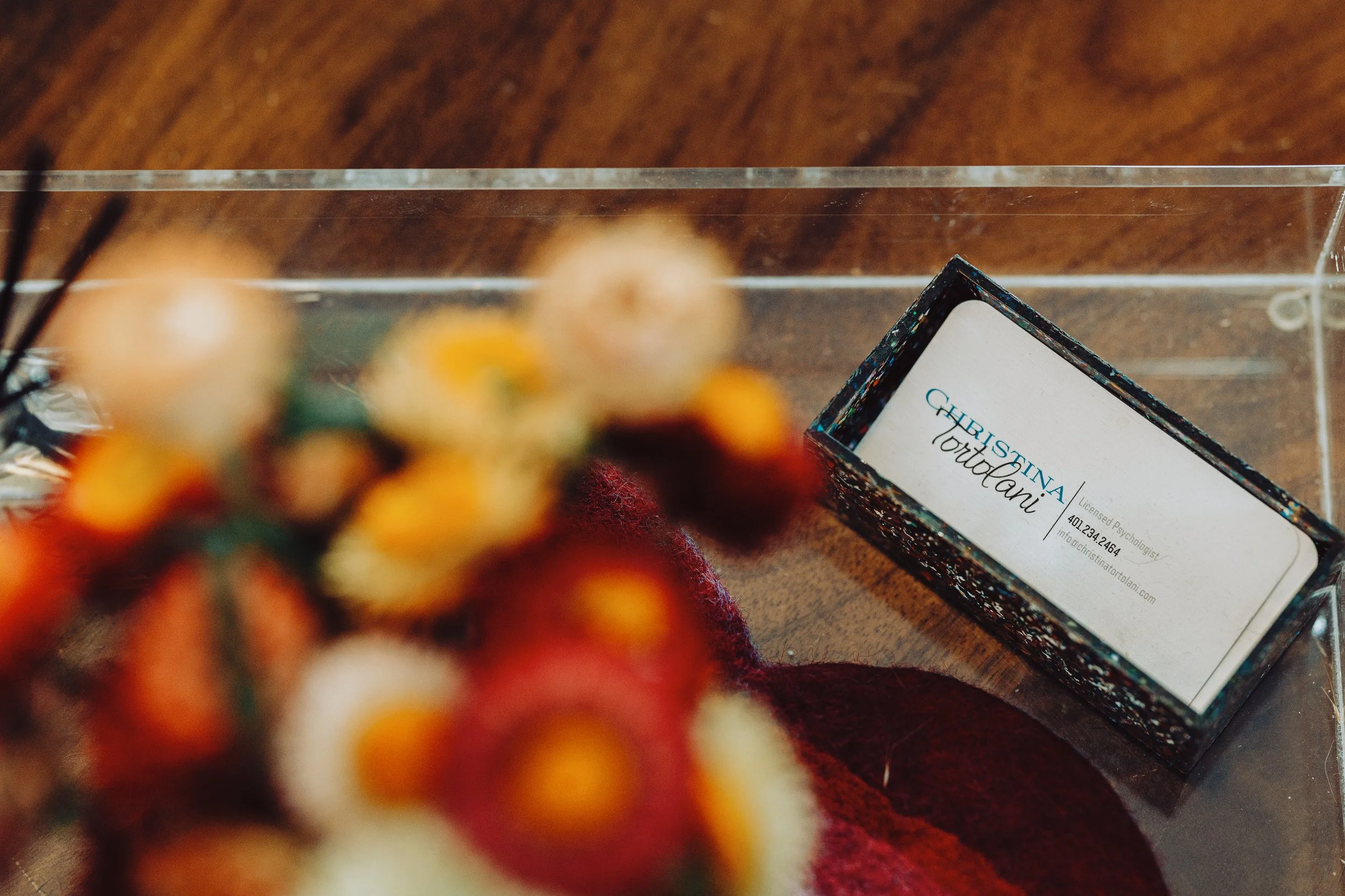 Close-up of a blurred mosaic display of colorful tiles or beads, with a business card for 'Christina Tortolani' angled nearby on a wooden surface.