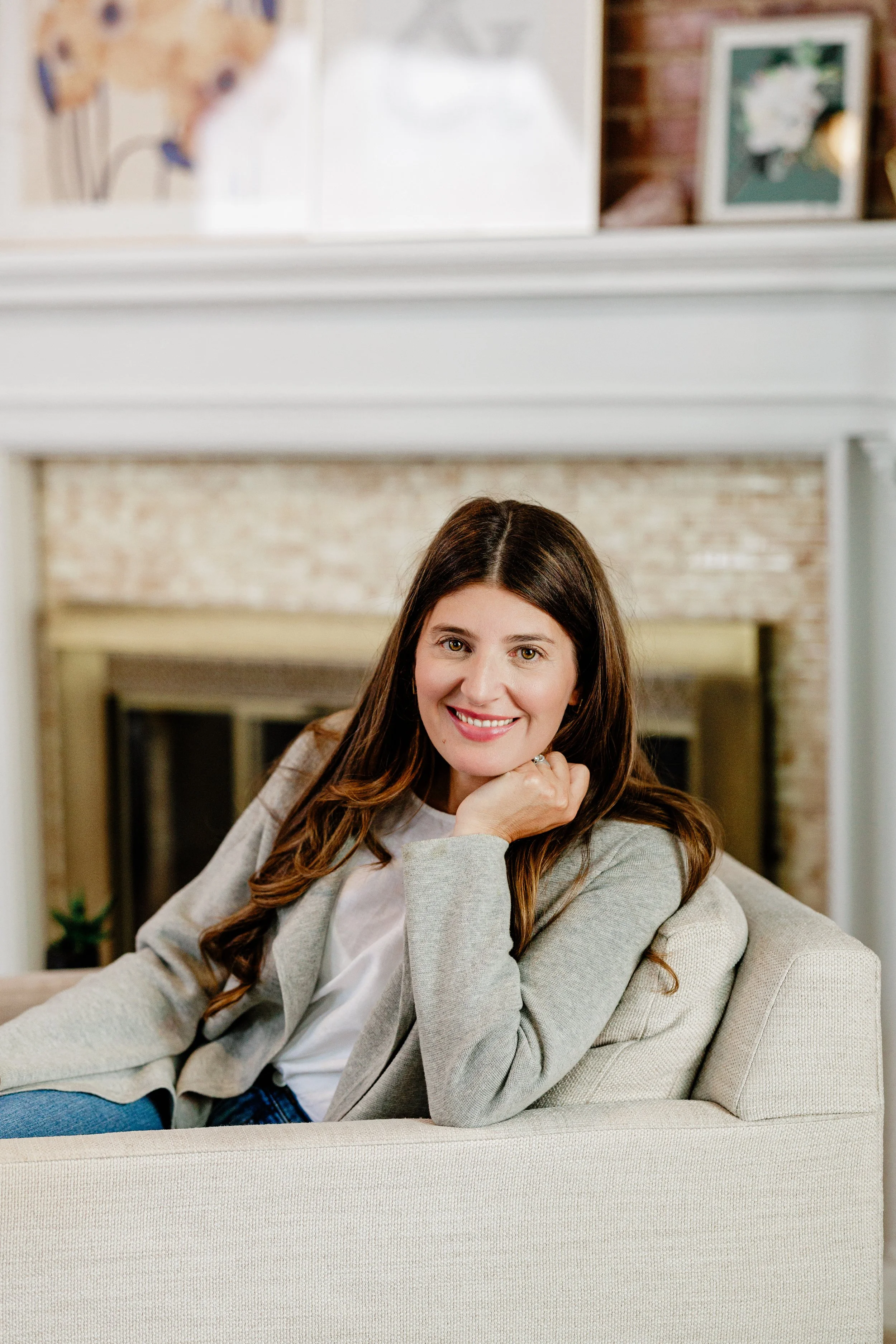 A woman with long brown hair smiling and sitting on a beige couch in a living room with a brick fireplace and framed photographs on the wall behind her.