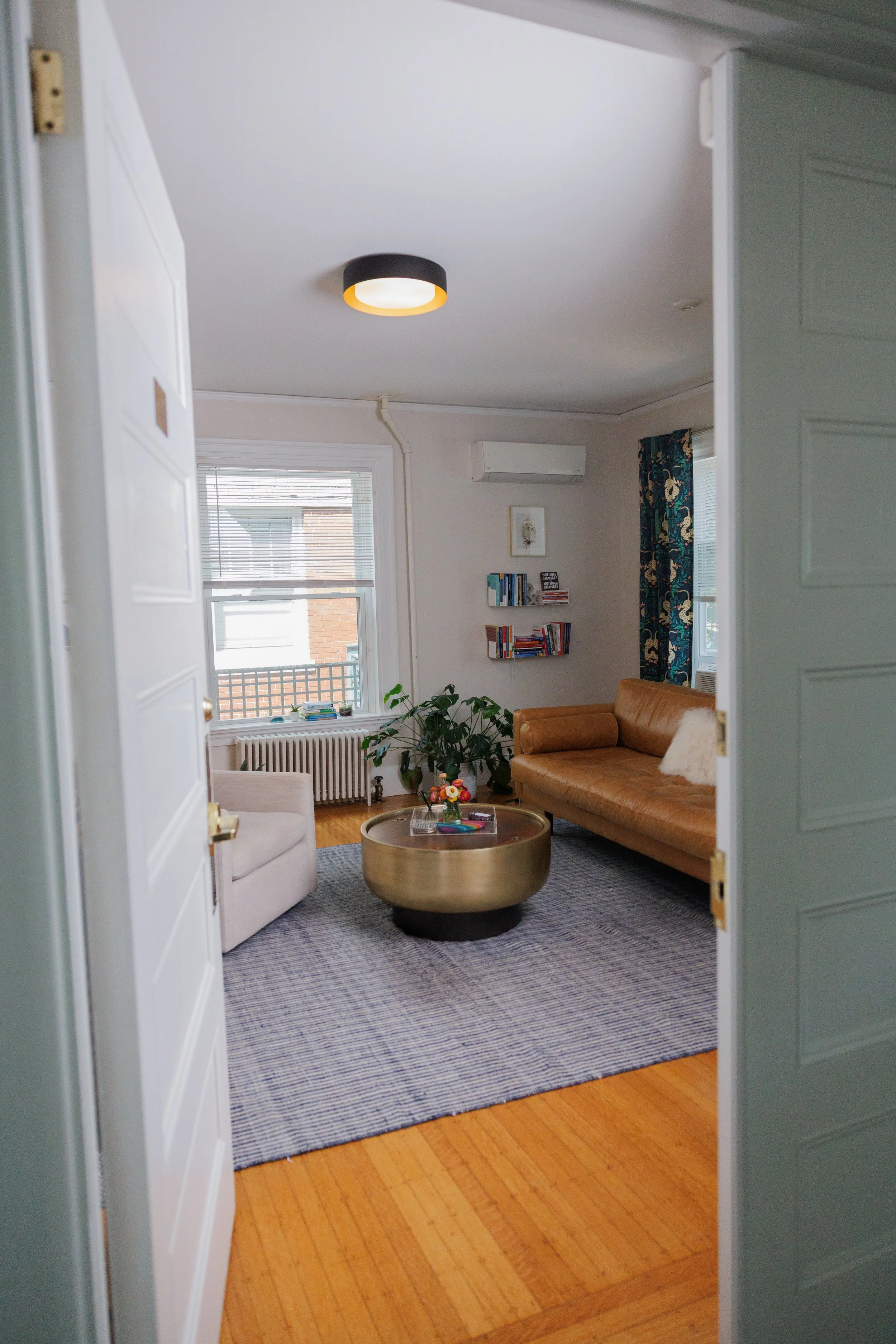 Living room with a brown sofa, beige armchair, small round coffee table, bookshelves, large window with blinds, sheer curtains with a floral pattern, potted plant, and air conditioning unit on the wall.