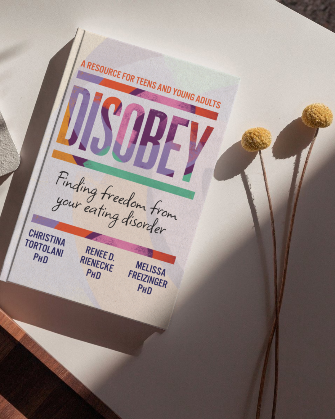 Cover of a book titled "Disobey: A Resource for Teens and Young Adults" with the subtitle "Finding freedom from your eating disorder," authored by Christina Tortolani PhD, Rene D. Rienecke PhD, and Melissa Freizinger PhD. The book is placed on a white surface, illuminated by natural sunlight, with two yellow billy ball flowers nearby.